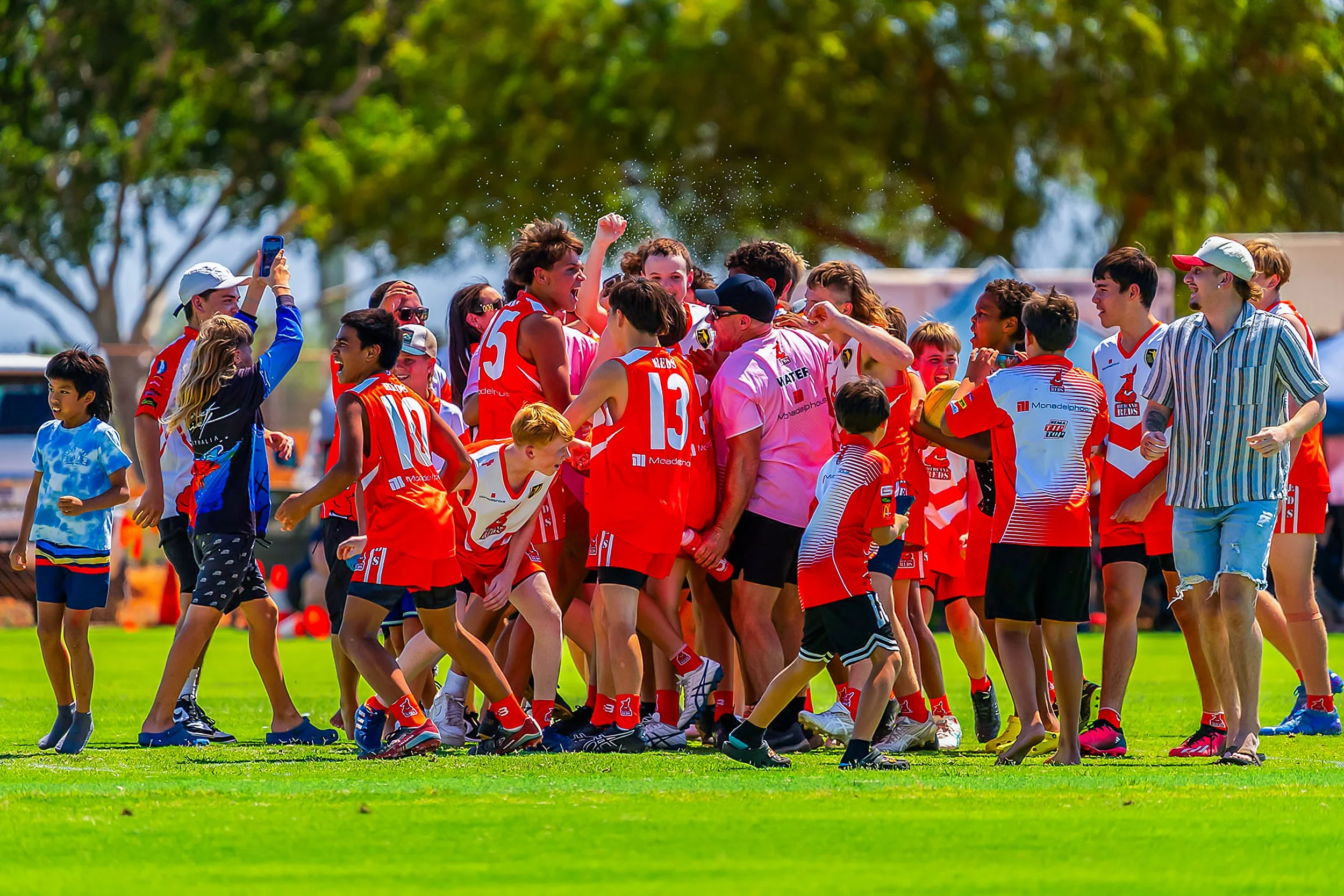 A group of children and adults celebrating on a sports field, wearing red sports uniforms, with some taking photos and others cheering.