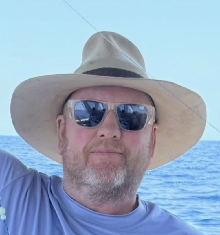 A man wearing a wide-brimmed hat and sunglasses taking a selfie on a boat with the ocean in the background.