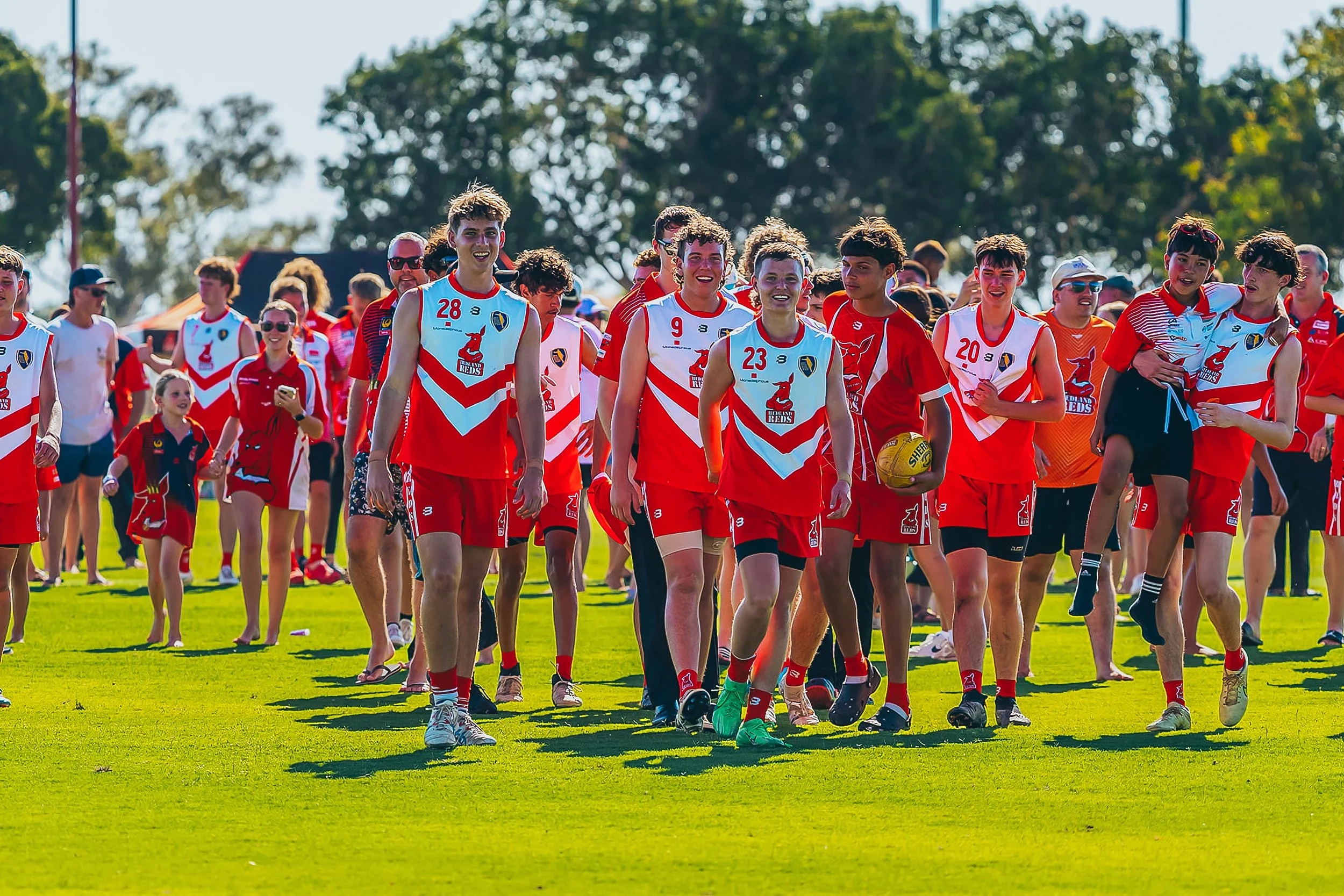 Group of young people, mostly boys, wearing red and white sports uniforms walking on a sunny grassy field after a sports game, with a background of trees and a clear blue sky.
