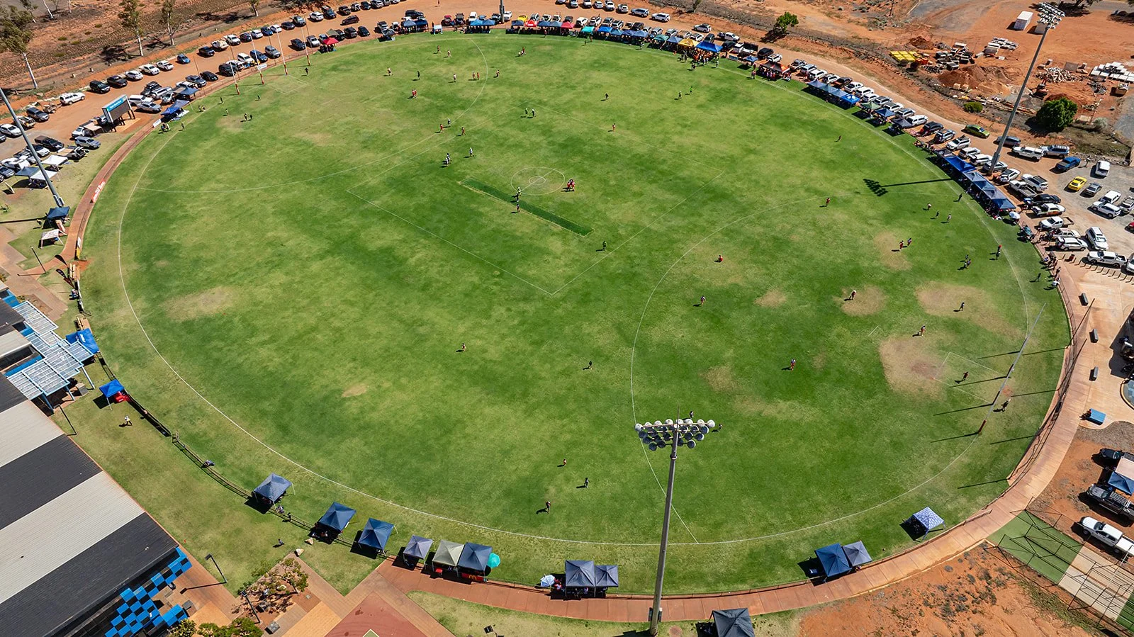 An aerial view of a large oval sports field with players playing what appears to be cricket, surrounded by cars parked along the perimeter and tents along the edge of the field.