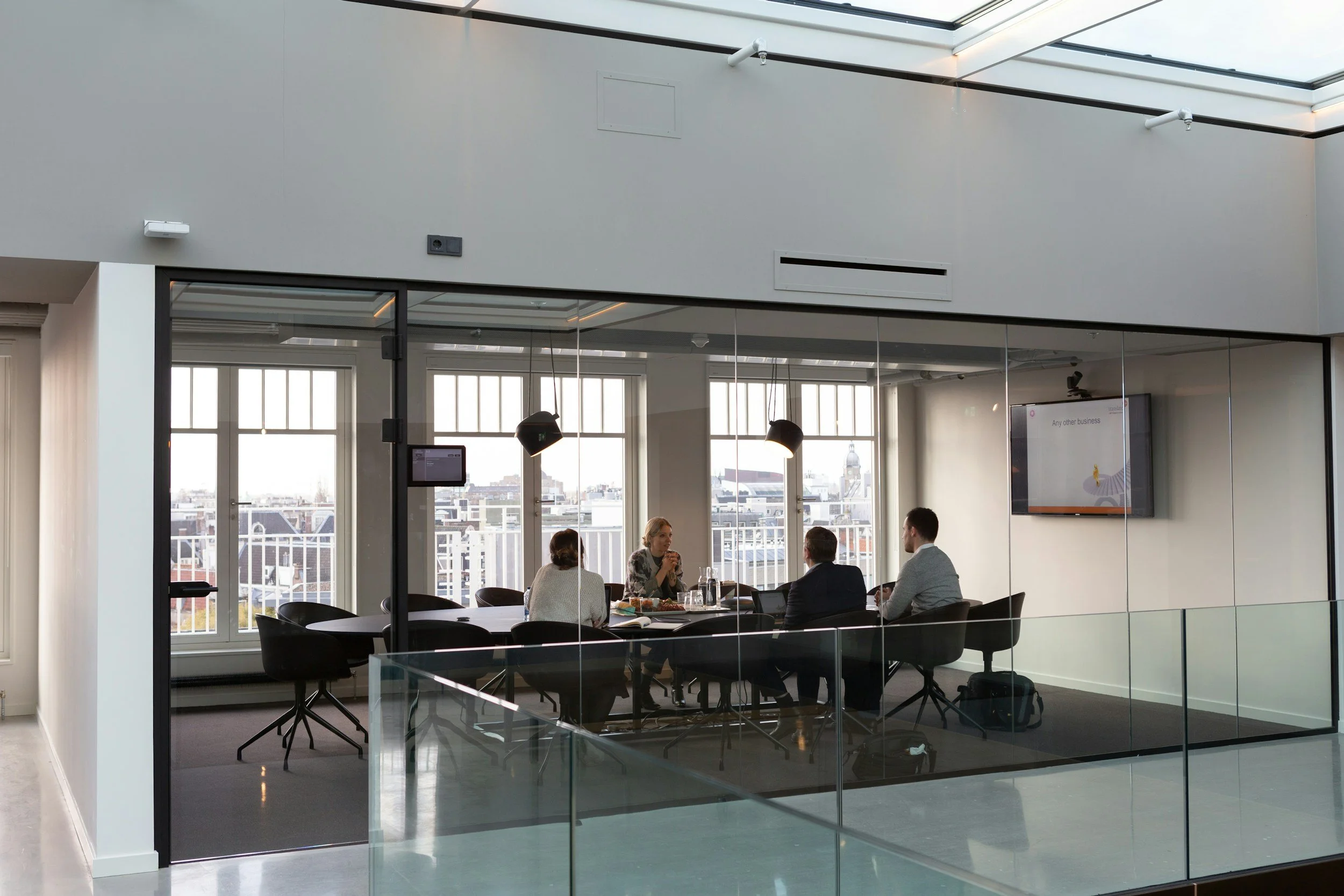 Business meeting with four people sitting around a table in a glass-walled conference room with city view and television screen.