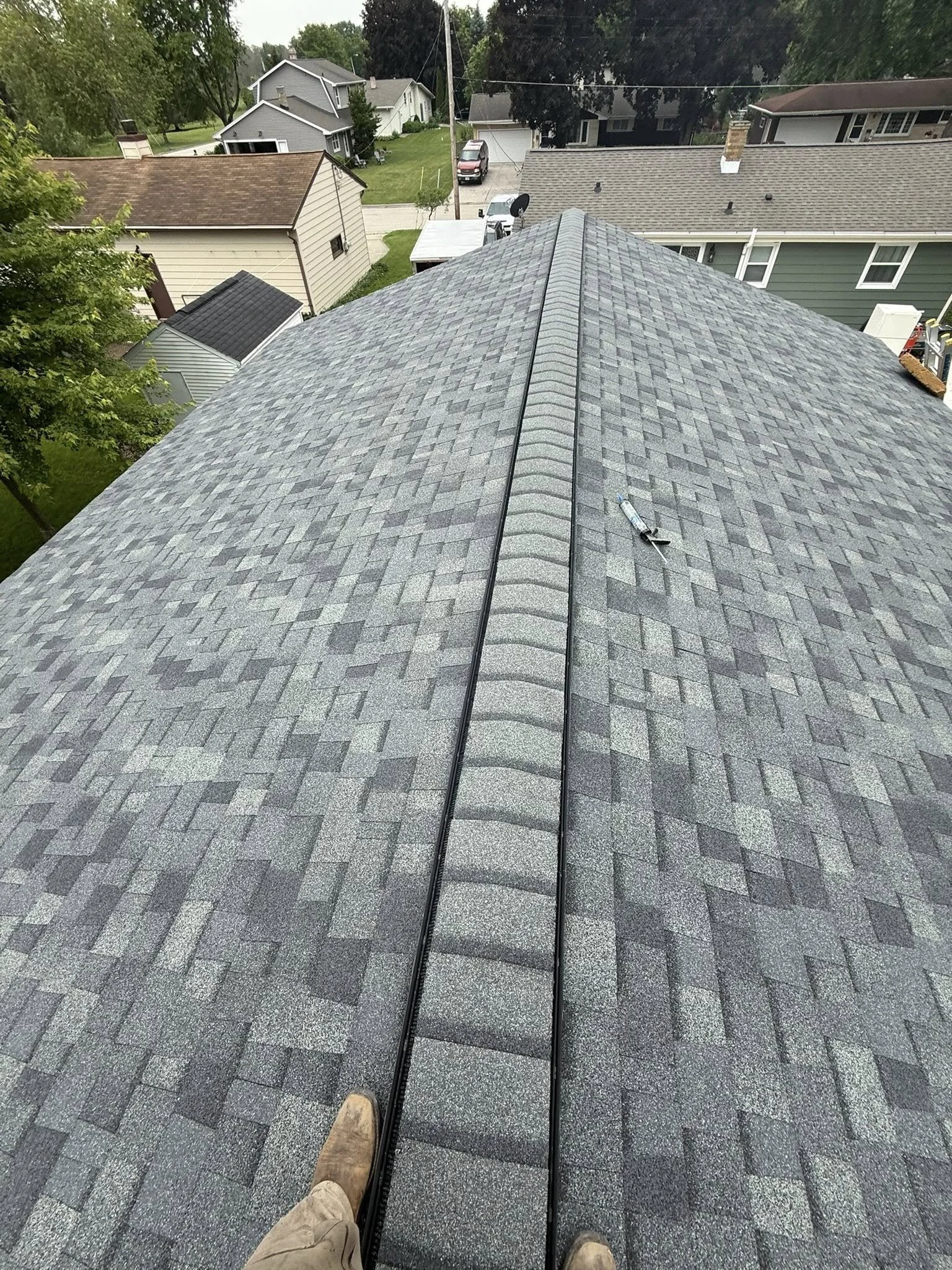 A view from the roof of a house showing grey asphalt shingles, a vent pipe on the roof, and neighboring houses and trees in the background.