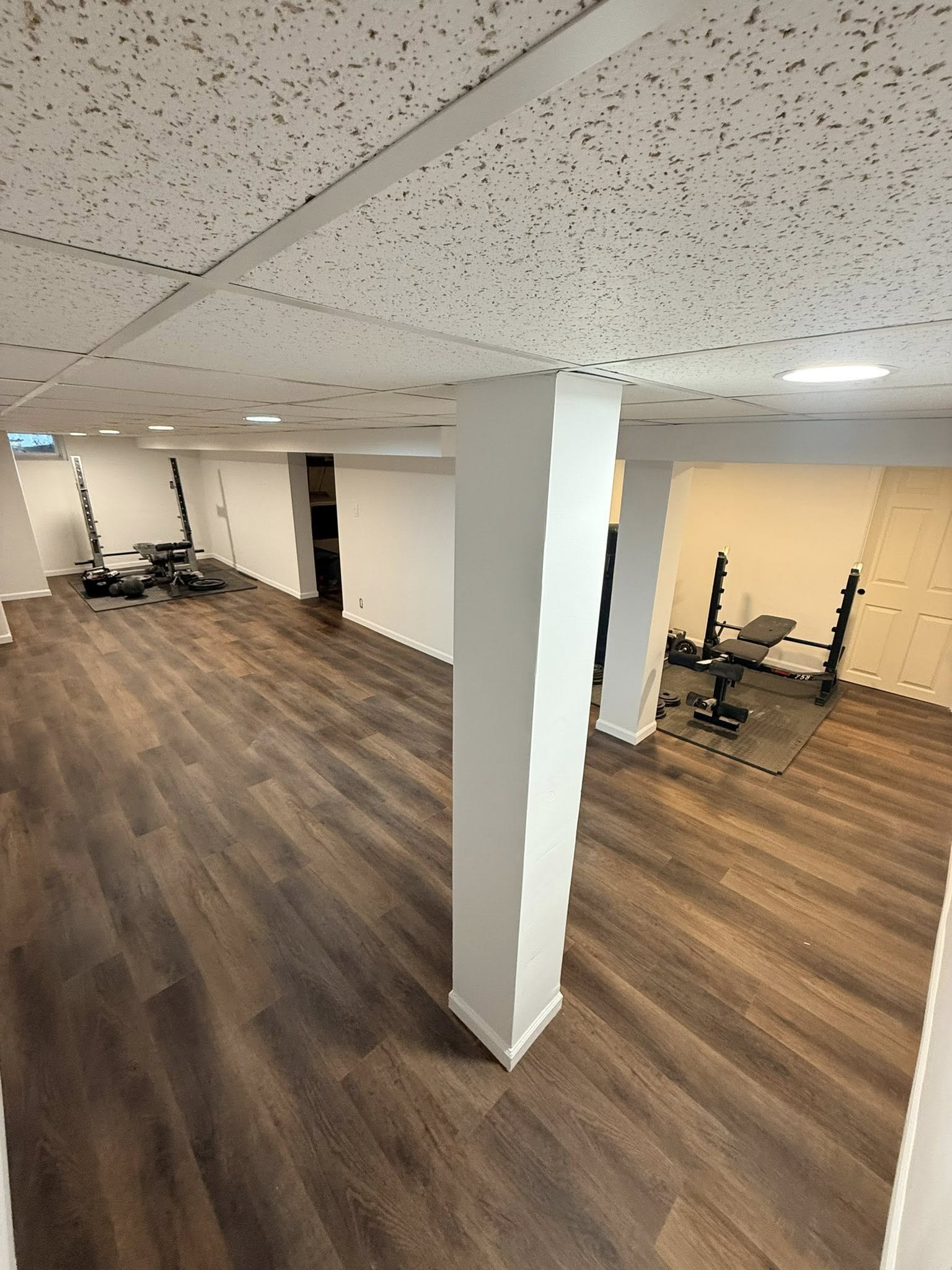 Finished basement with wood-look flooring, white walls, ceiling tiles, and workout equipment including a squat rack, bench, and weights.
