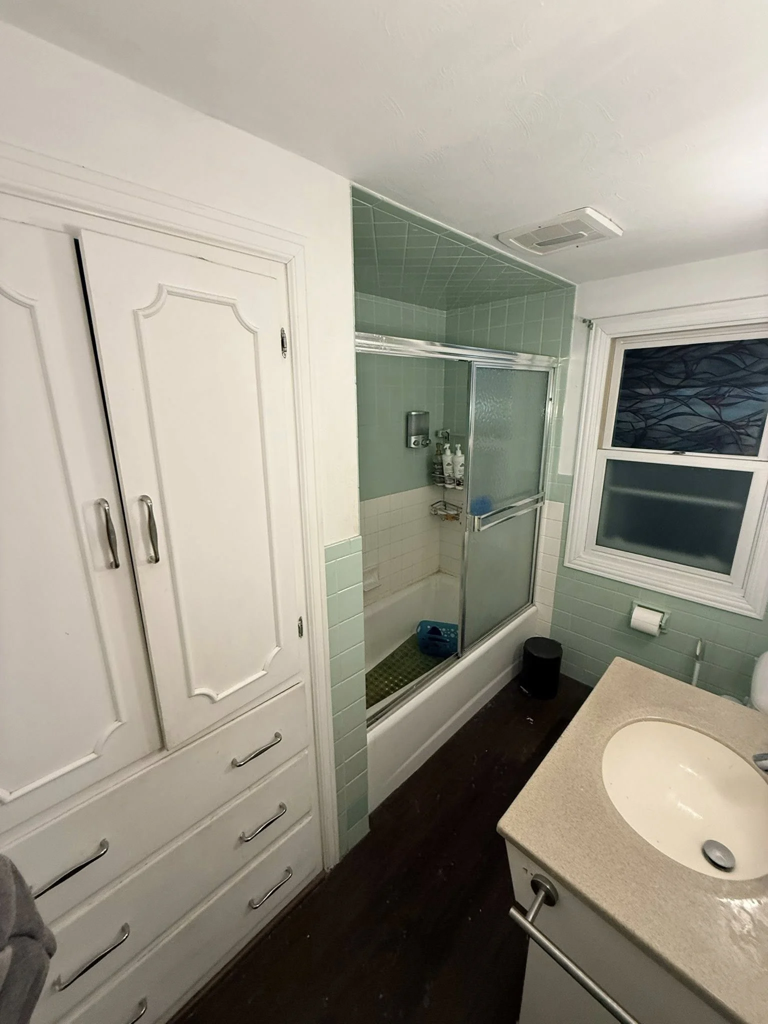 Bathroom with a closed white cabinet, a shower with green tiles and frosted glass sliding doors, a sink with a beige countertop, a window, and a toilet paper holder.