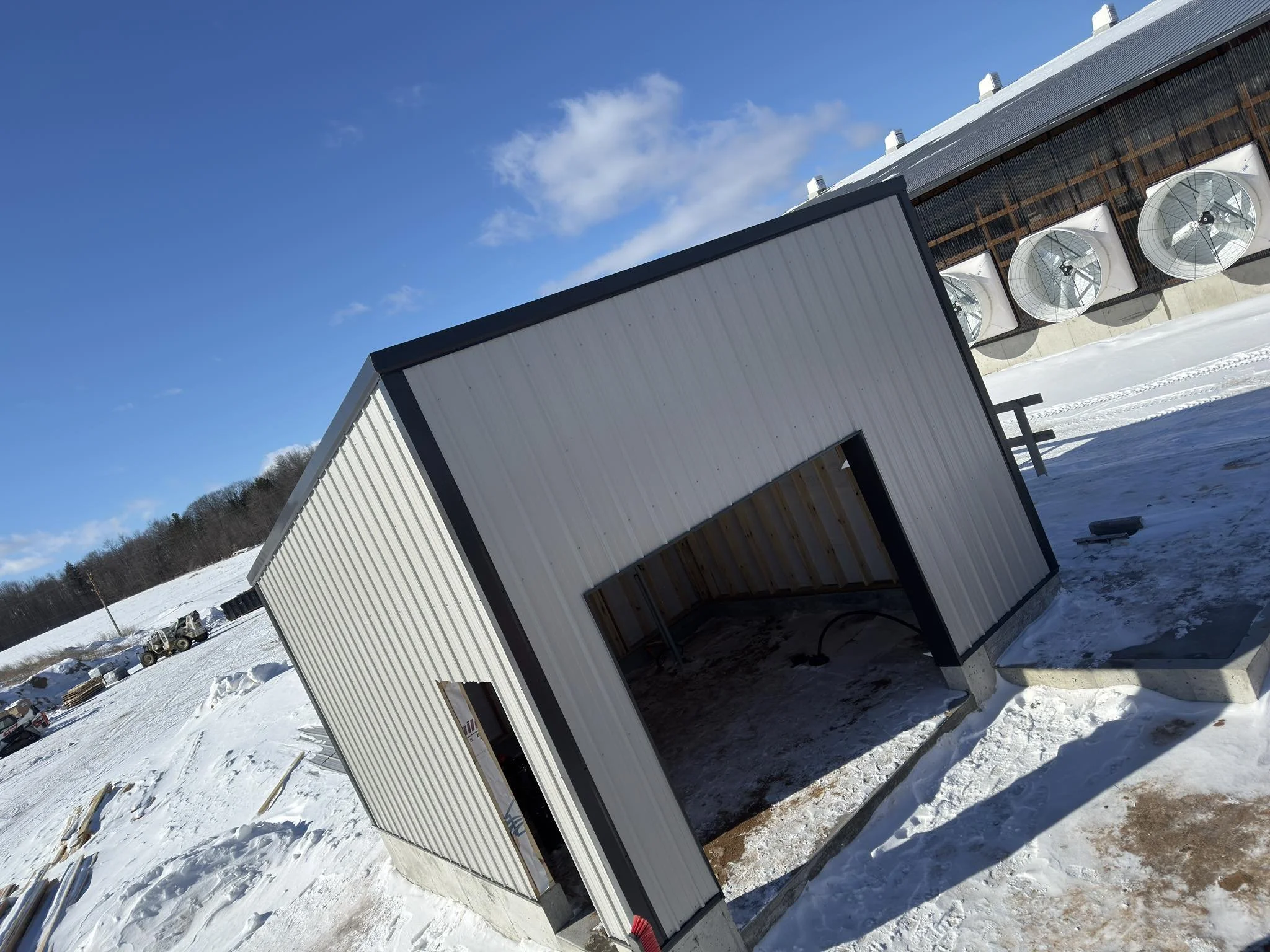 An outdoor construction site in winter with a metal shed and a building with large exhaust fans. The ground is covered in snow and the sky is clear with some clouds.