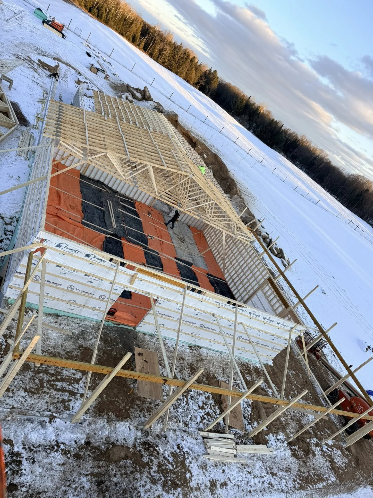 Construction site in winter showing the framework of a building being erected, with wooden beams, partially assembled walls, and snow on the ground and surrounding area.