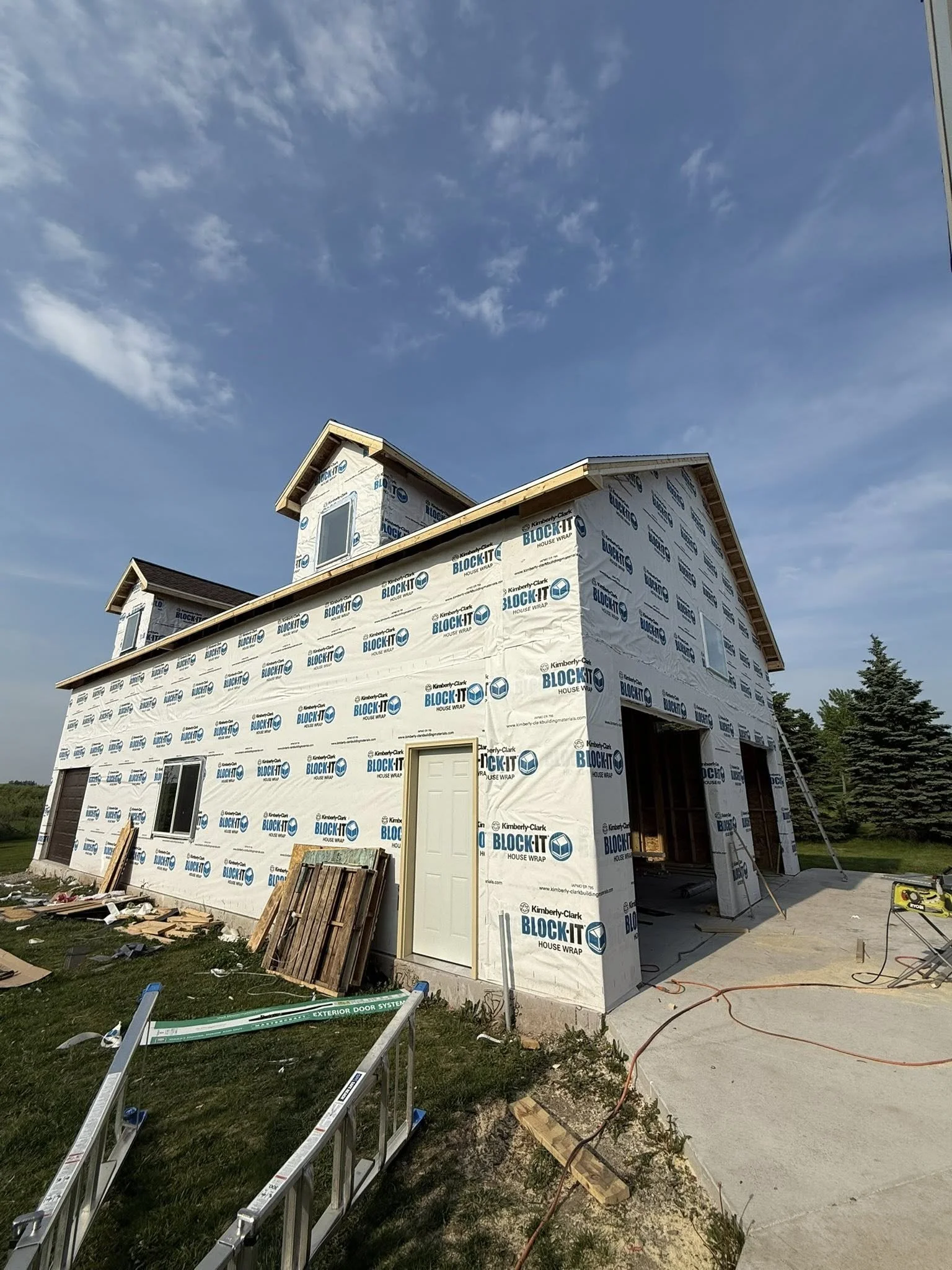 A house under construction, wrapped in house wrap with windows installed, a ladder leaning against the side, and construction tools scattered around.