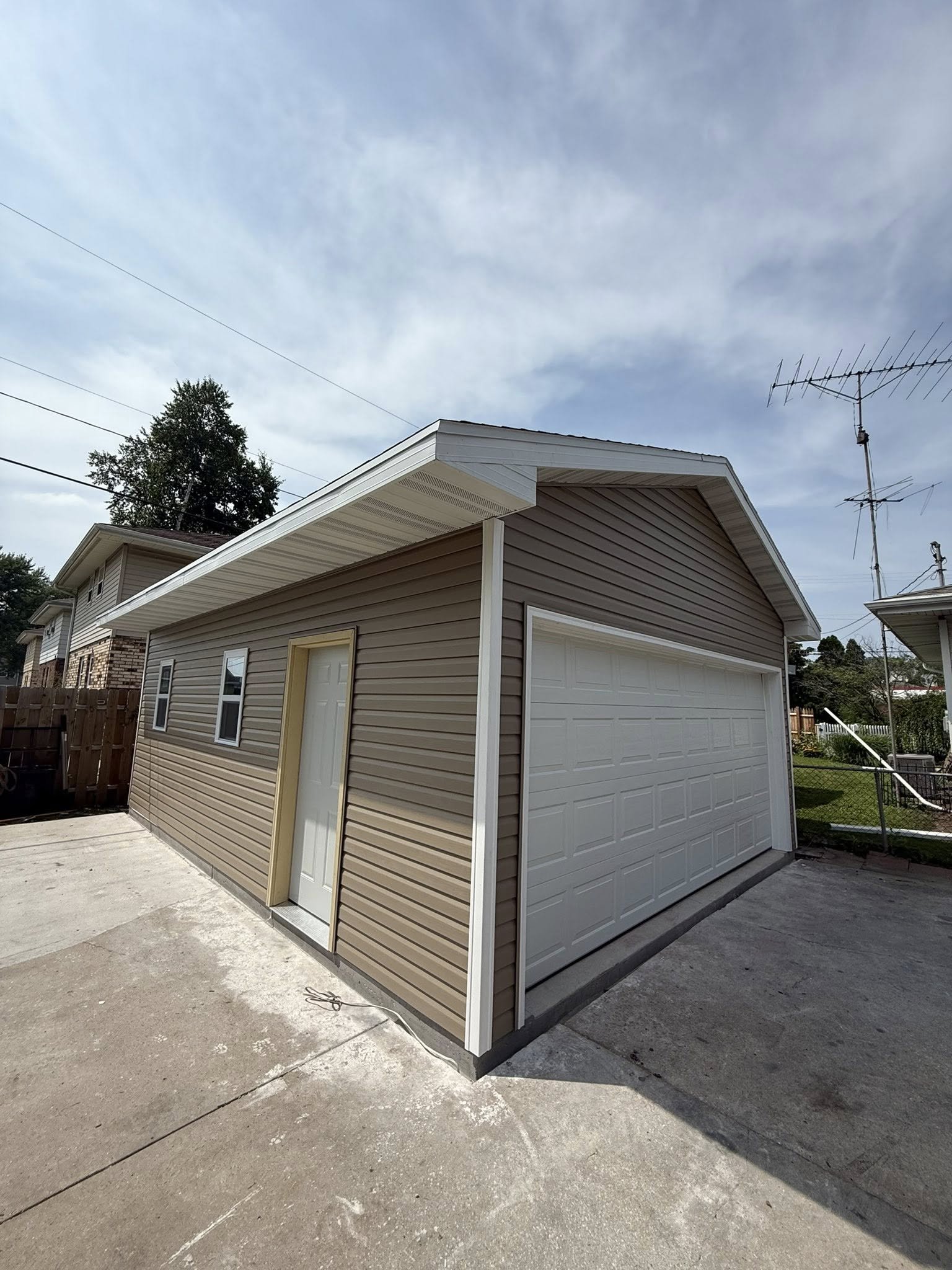 A beige detached garage with a rolling door and a side door, situated on a concrete driveway in a residential area with neighboring houses and a fence, under a partly cloudy sky.