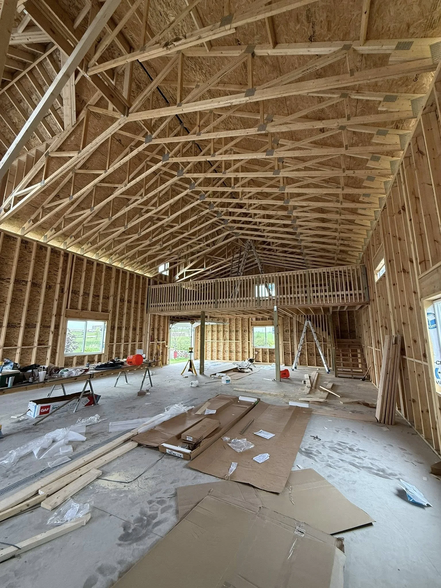 The interior of a large building under construction with exposed wooden framing, high vaulted ceiling, and a second-floor balcony. Construction materials and tools are scattered across the unfinished concrete floor.