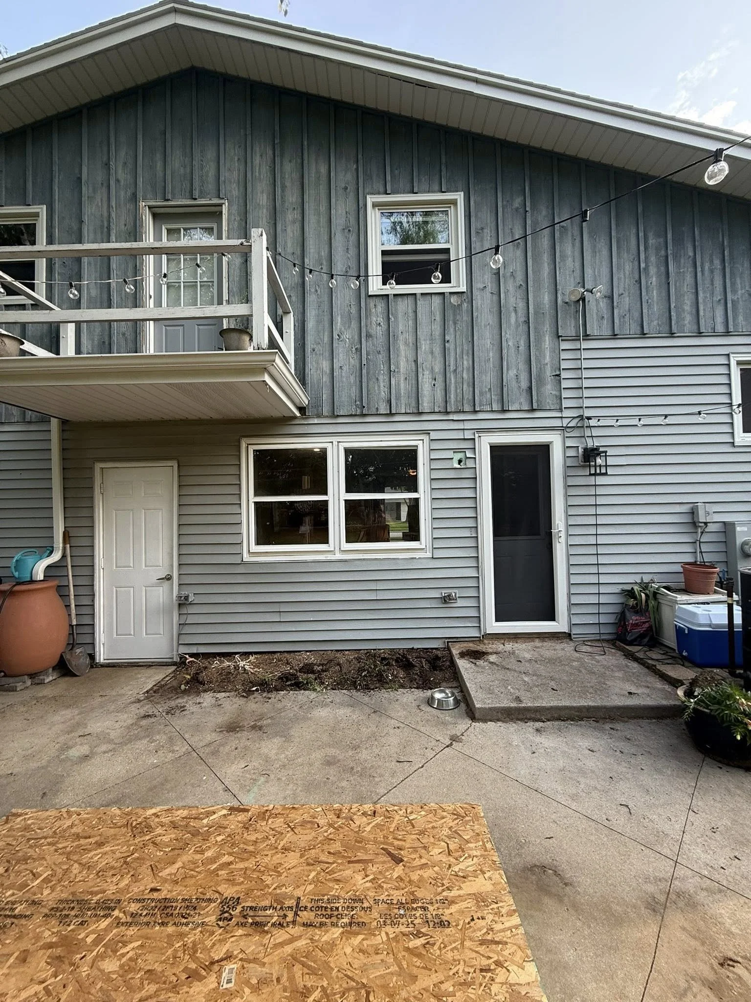 Backyard view of a house with a lower level in gray vinyl siding and an upper level with weathered wood paneling. There is a small second-floor balcony with a railing and pots, a string of outdoor lights, two windows on the upper level, and two doors on the lower level. The ground is concrete with some dirt and construction materials, including a piece of plywood at the front.