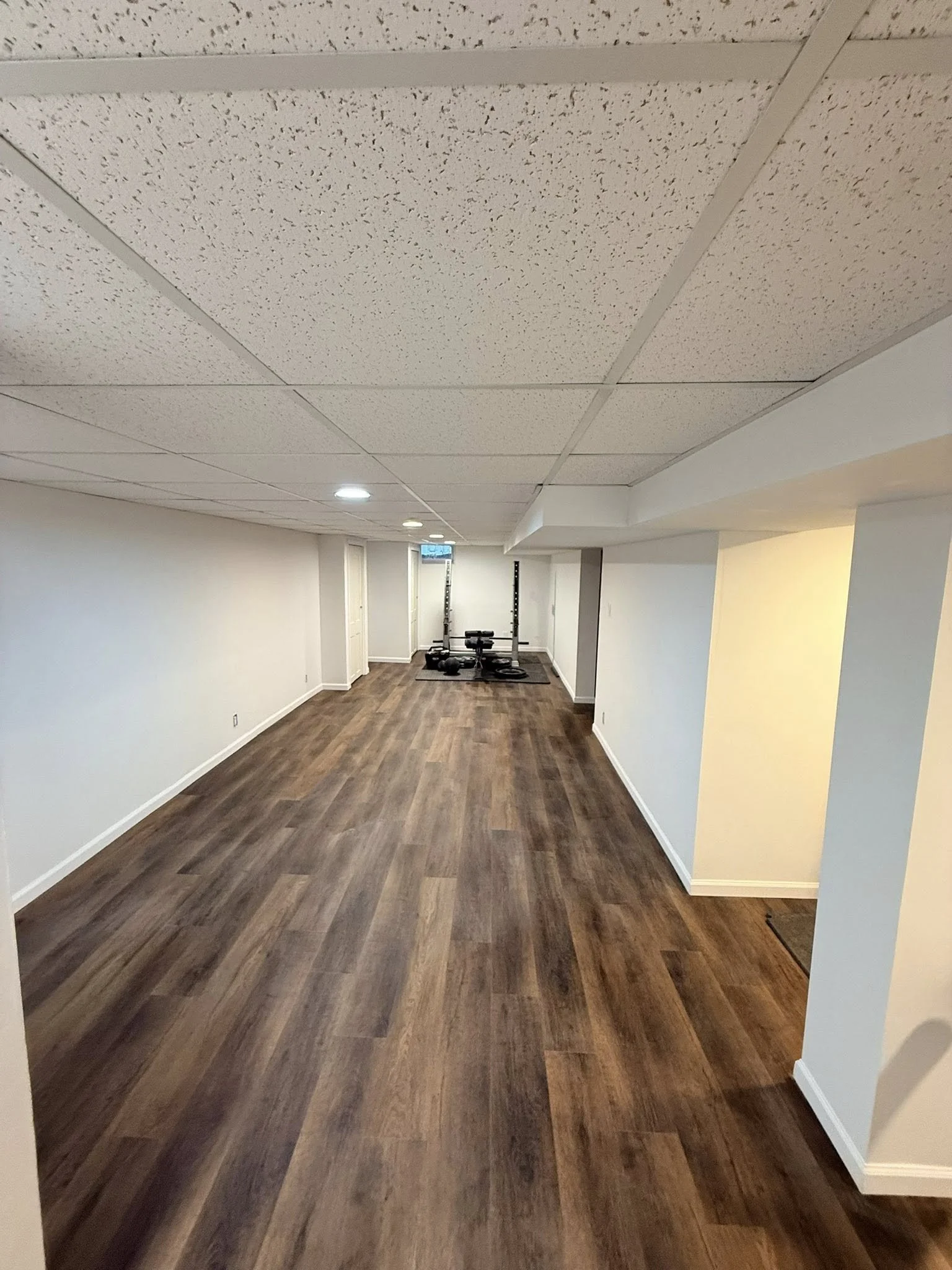 Empty basement room with wood flooring, white walls, and a small workout station at the far end.