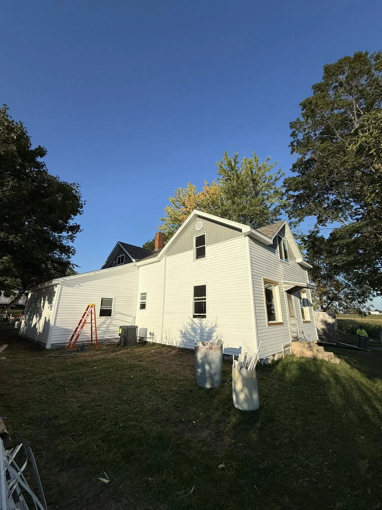 Side view of a white two-story house under construction with an orange ladder, trash bins, and construction workers nearby, surrounded by green trees and under a clear blue sky.
