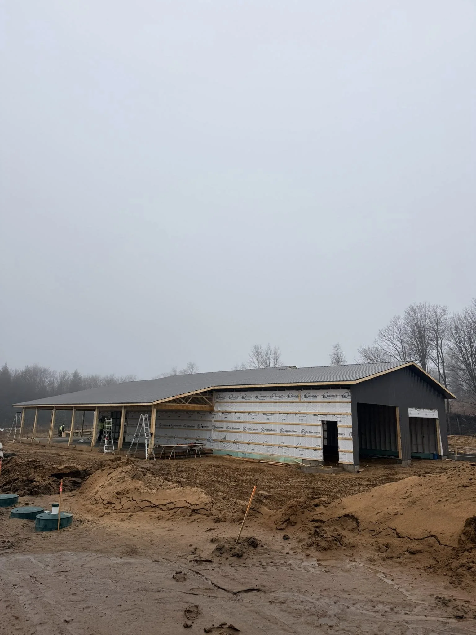 Construction site with a partially built barn under construction, with construction materials and equipment around, and trees in the background.