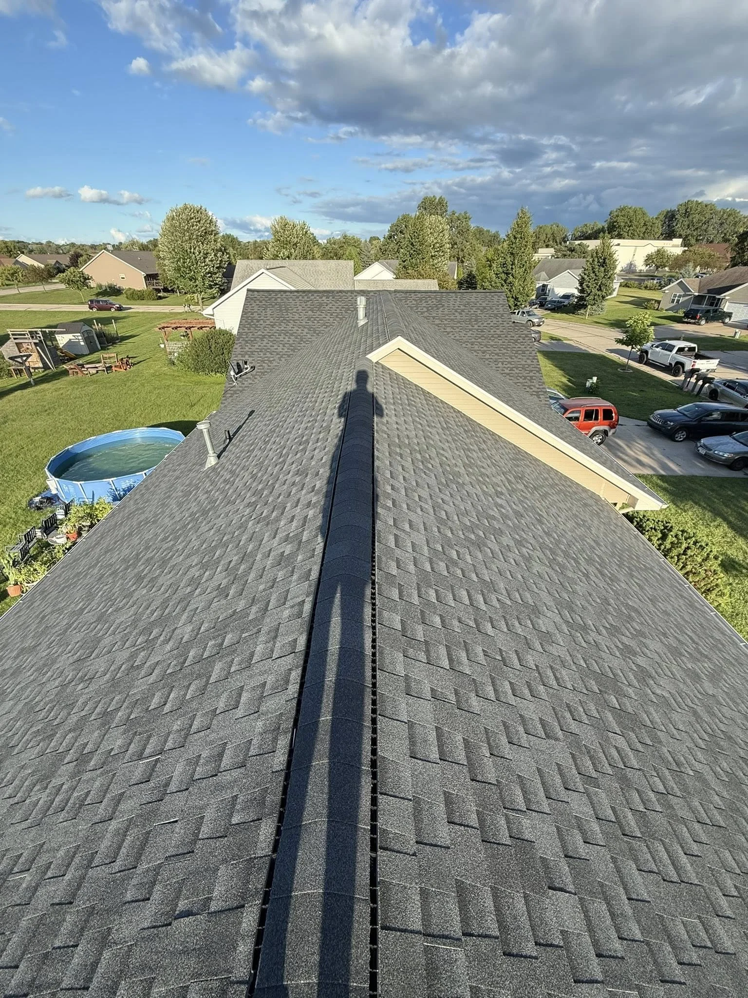 View from the roof of a suburban neighborhood with houses, cars, trees, and a blue sky with clouds.