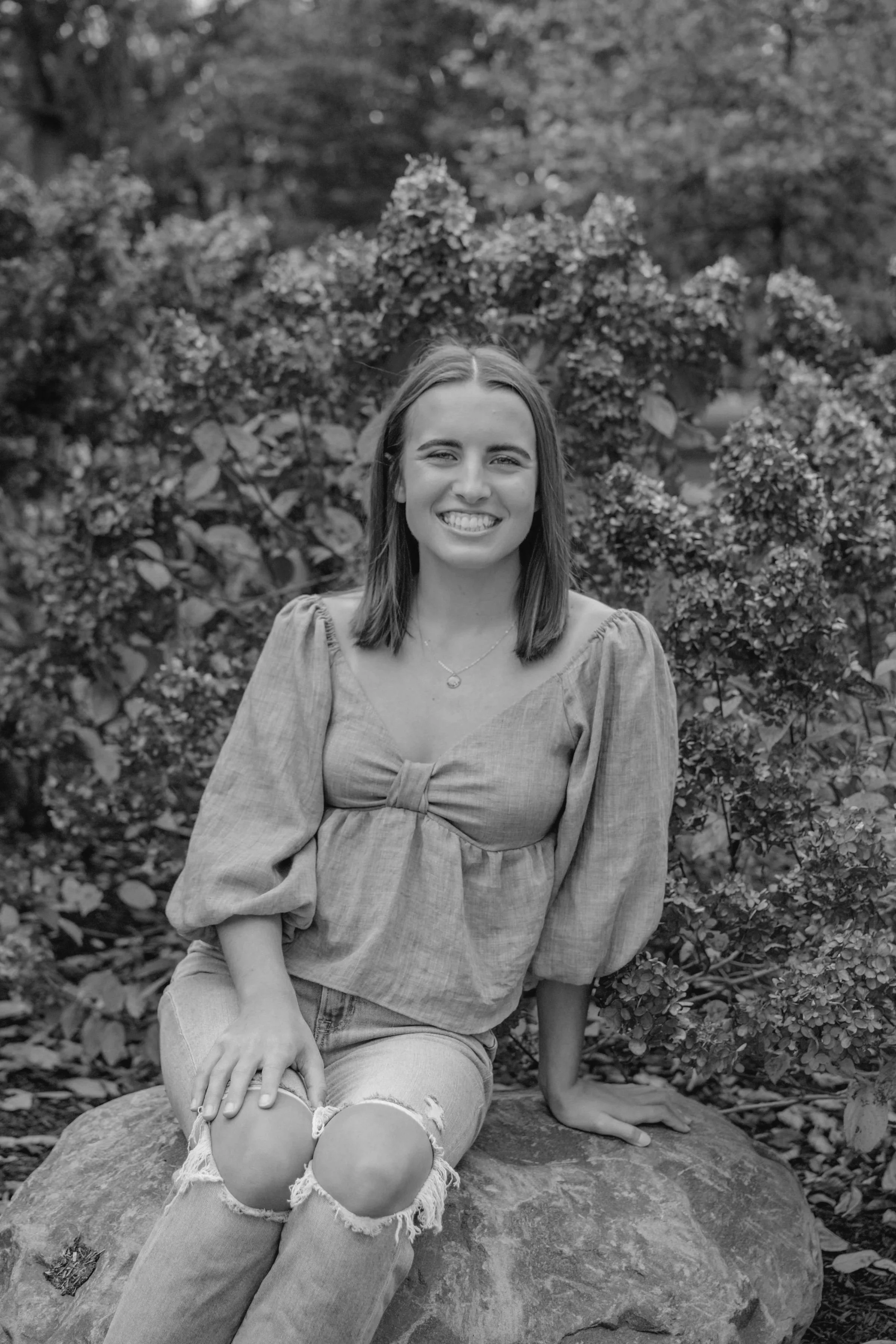 A young woman with shoulder-length hair smiling, sitting on a large rock outdoors, surrounded by bushes and trees.