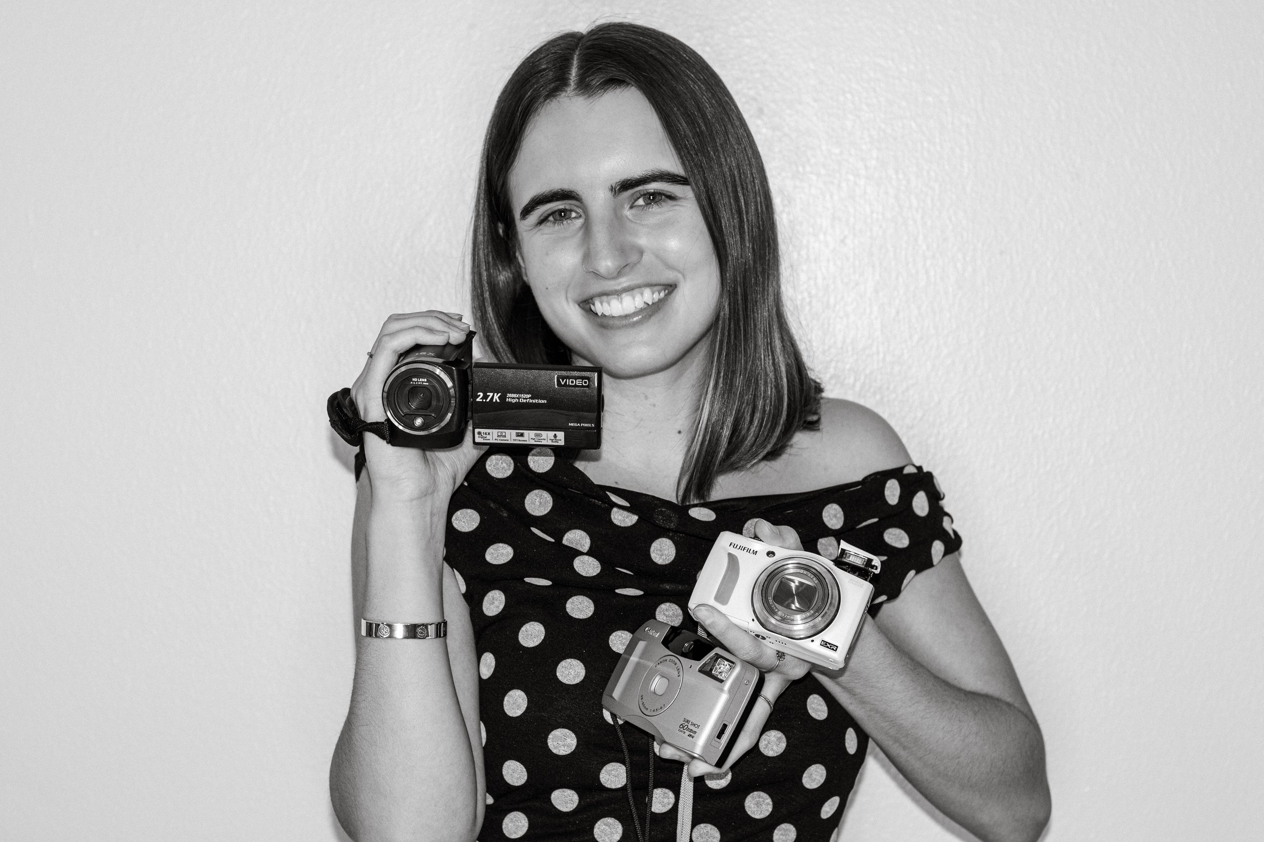 A smiling woman holding three different cameras, wearing a polka dot top, against a plain wall.