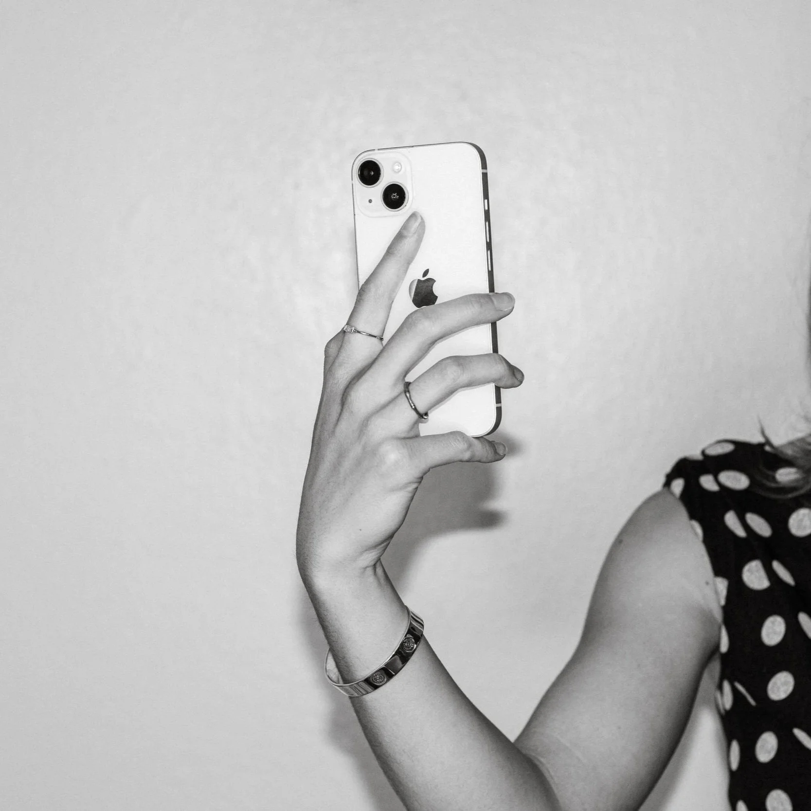 A person holding an iPhone taking a selfie, with only their arm and part of their shoulder visible, against a plain background.
