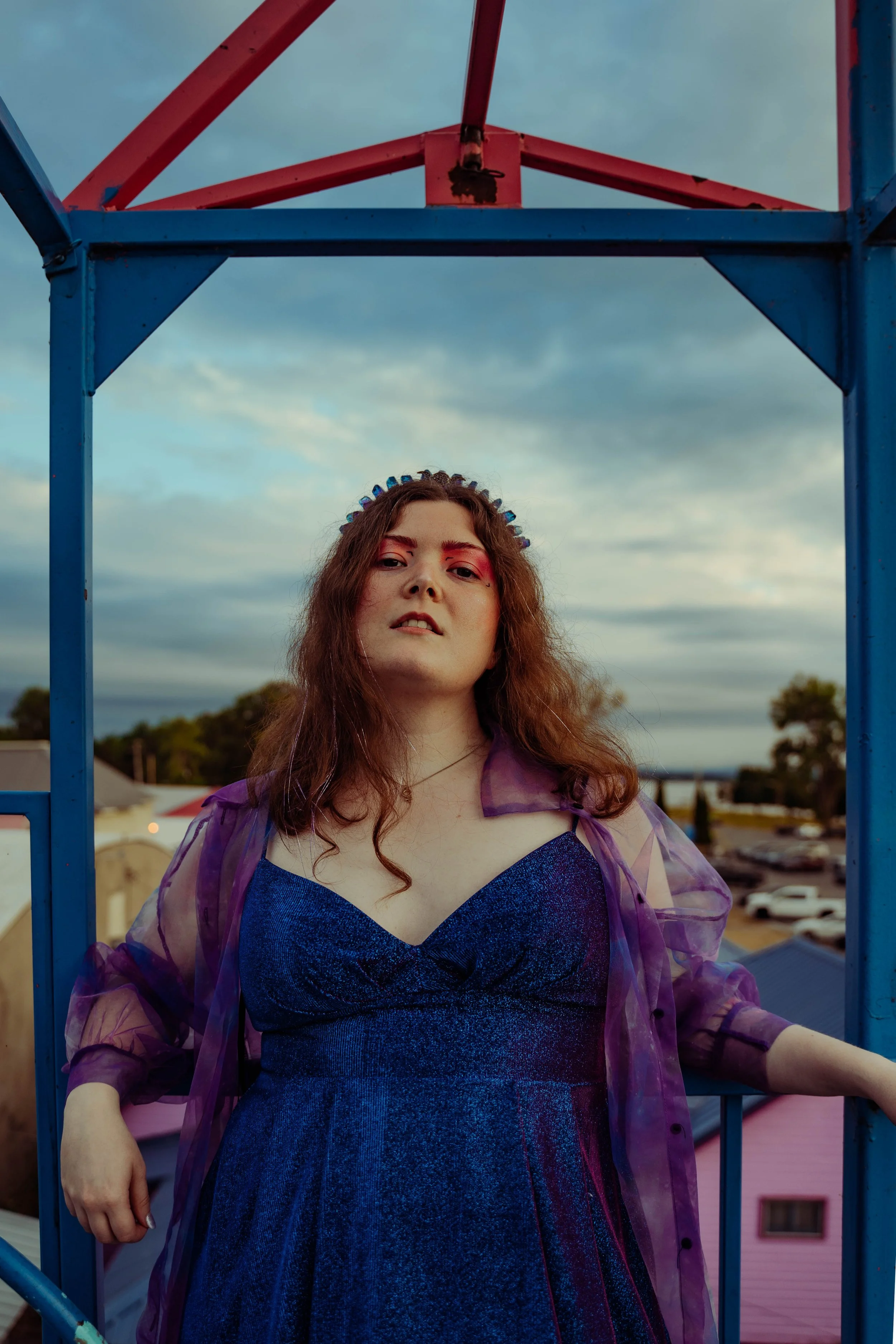 A young woman with wavy brown hair, wearing a sparkly blue dress and a sheer purple cover-up, standing on a colorful amusement park ride with a cloudy sky in the background.