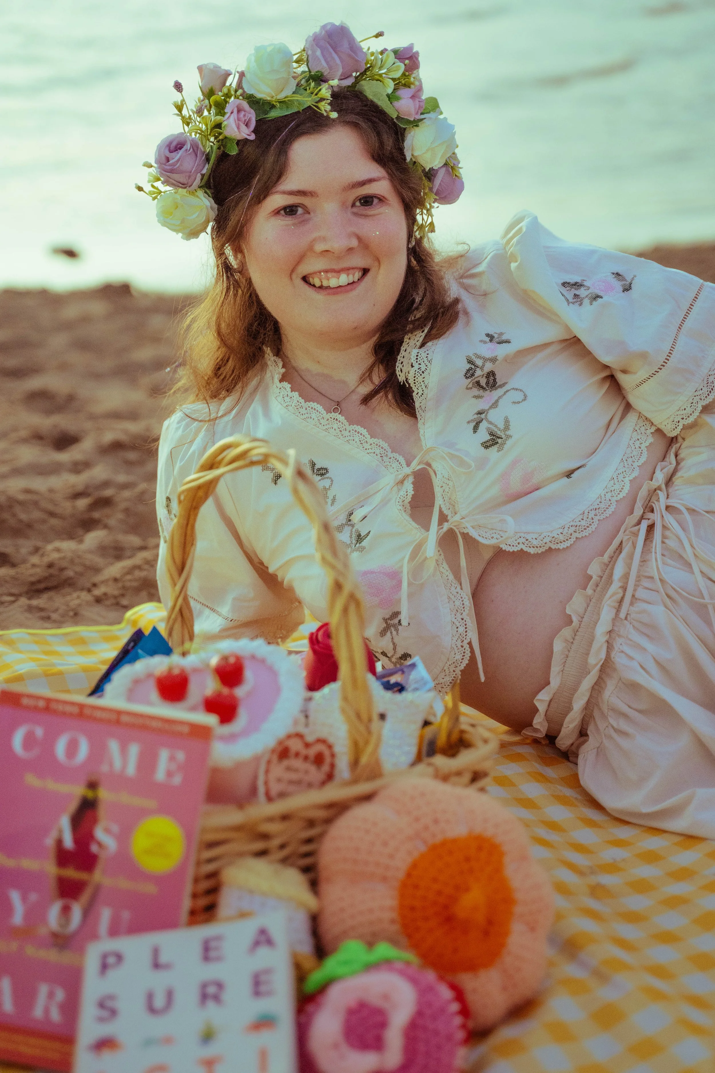 A young woman lying on a checkered picnic blanket at the beach, wearing a white blouse and skirt with floral embroidery and a flower crown, smiling at the camera with a basket of colorful knitted toys and books about sex in front of her.