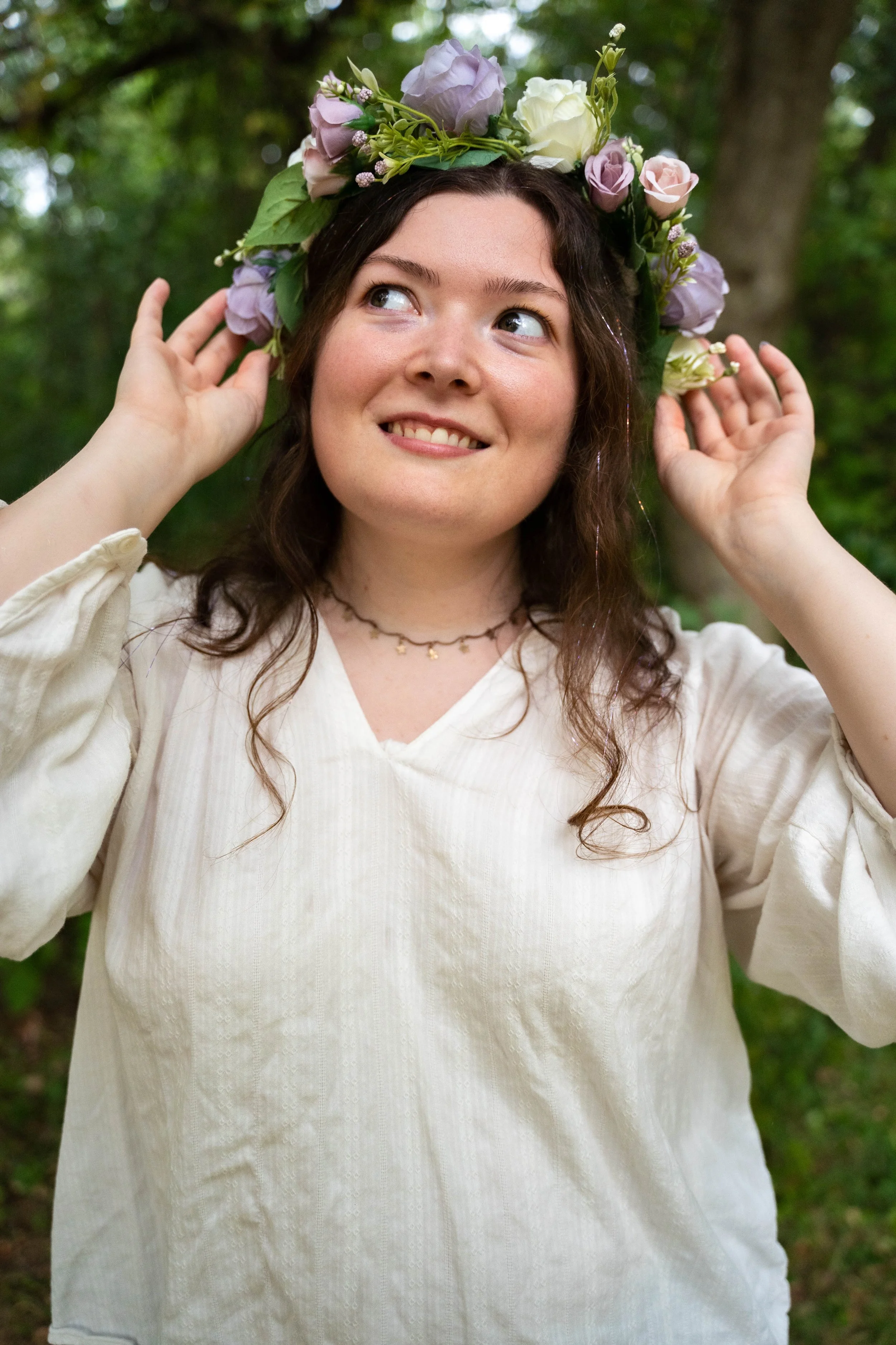 A young woman with wavy brown hair wearing a cream-colored blouse and a flower crown made of purple, white, and pink flowers standing outdoors in a green wooded area.