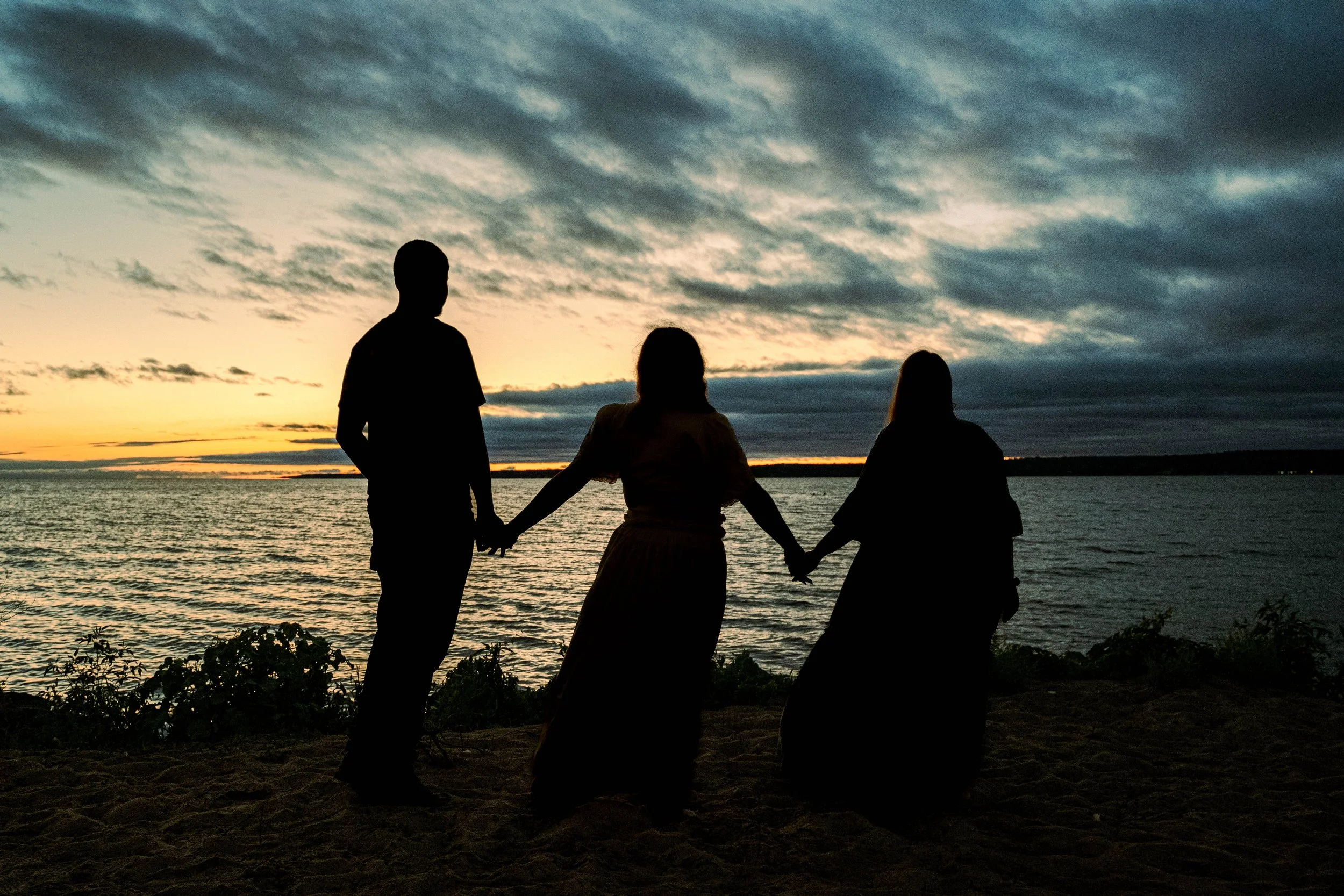 Three people holding hands by the water at sunset, silhouetted against a cloudy sky.