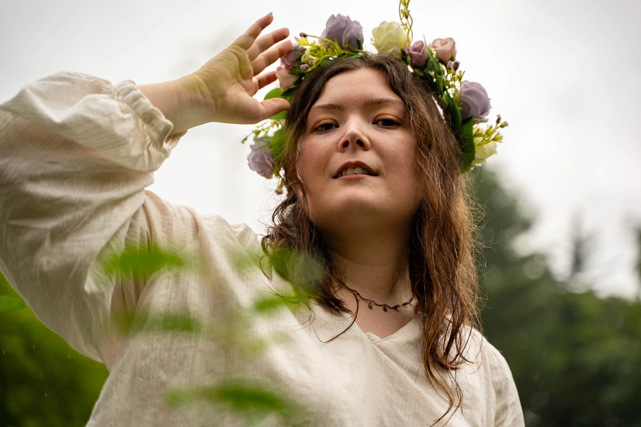 A young woman with wavy brown hair wearing a flower crown made of pink, purple, and white flowers, dressed in a cream-colored blouse, standing outdoors with blurred greenery in the background.