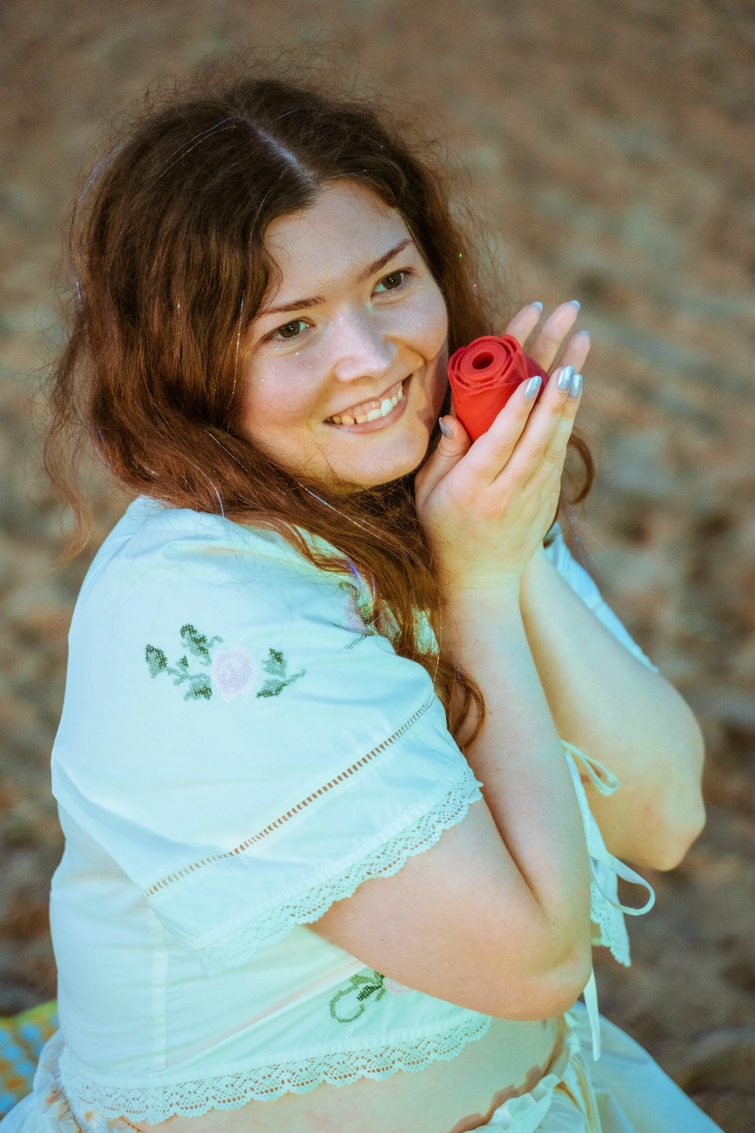 A young woman with wavy brown hair smiling and holding a red rose vibrator, sitting on a sandy beach wearing a white embroidered blouse with lace accents.