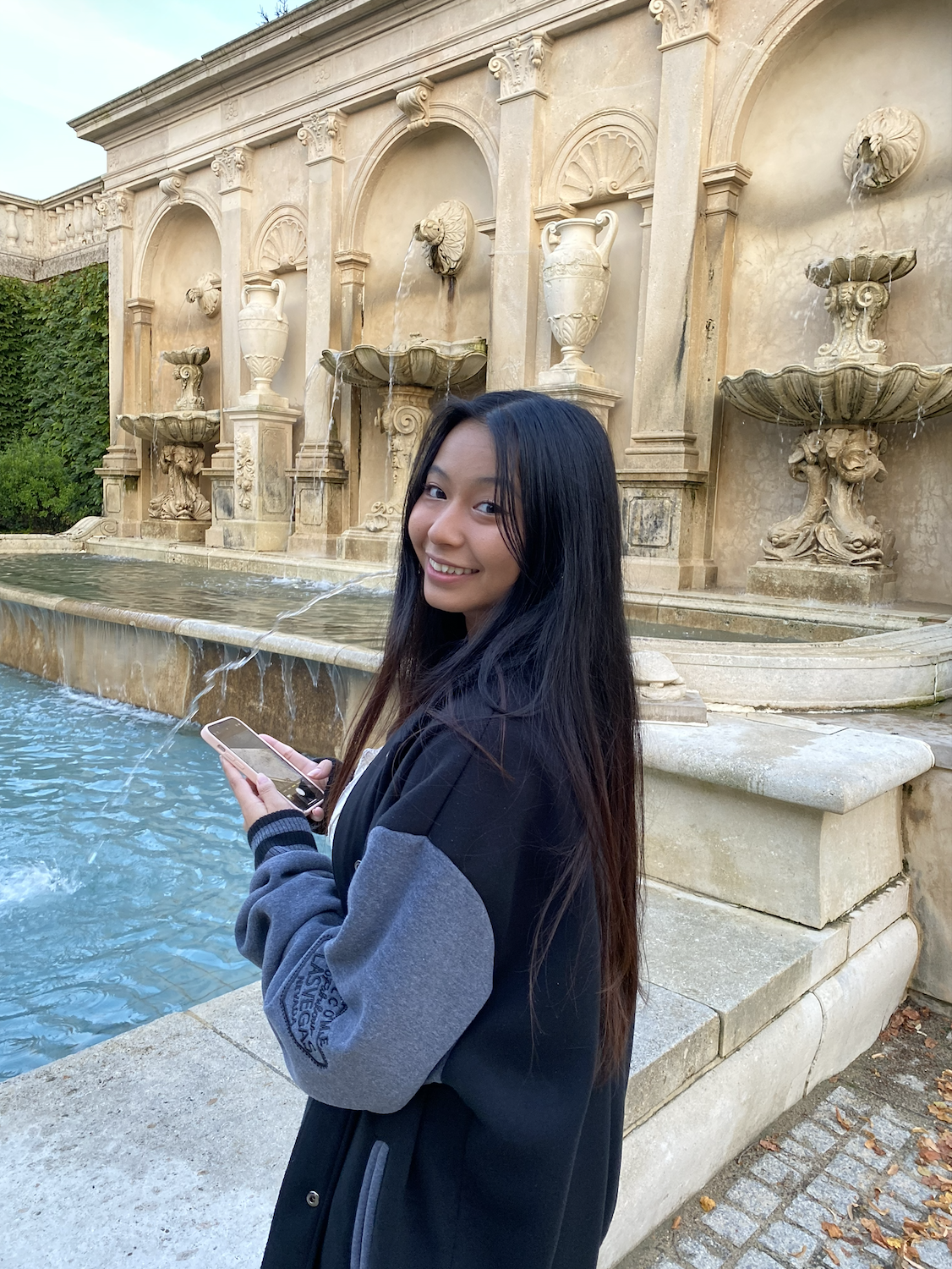 A young woman with long dark hair smiling while holding a phone, standing near a decorative fountain with water flowing from lion head spouts in an outdoor garden setting.