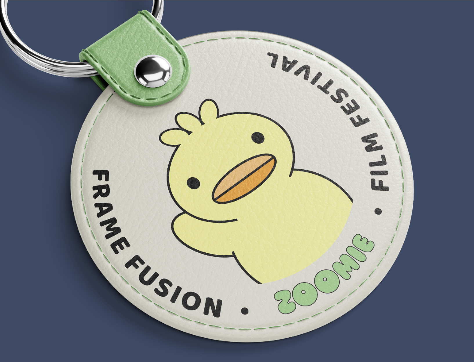 A circular merchandise tag with a cartoon yellow duck illustration in the center. The tag has text around the duck, including the phrase 'FRAMÉ FUSION,' and features a green strap with a metallic button, attached to a keyring. The background is dark blue.