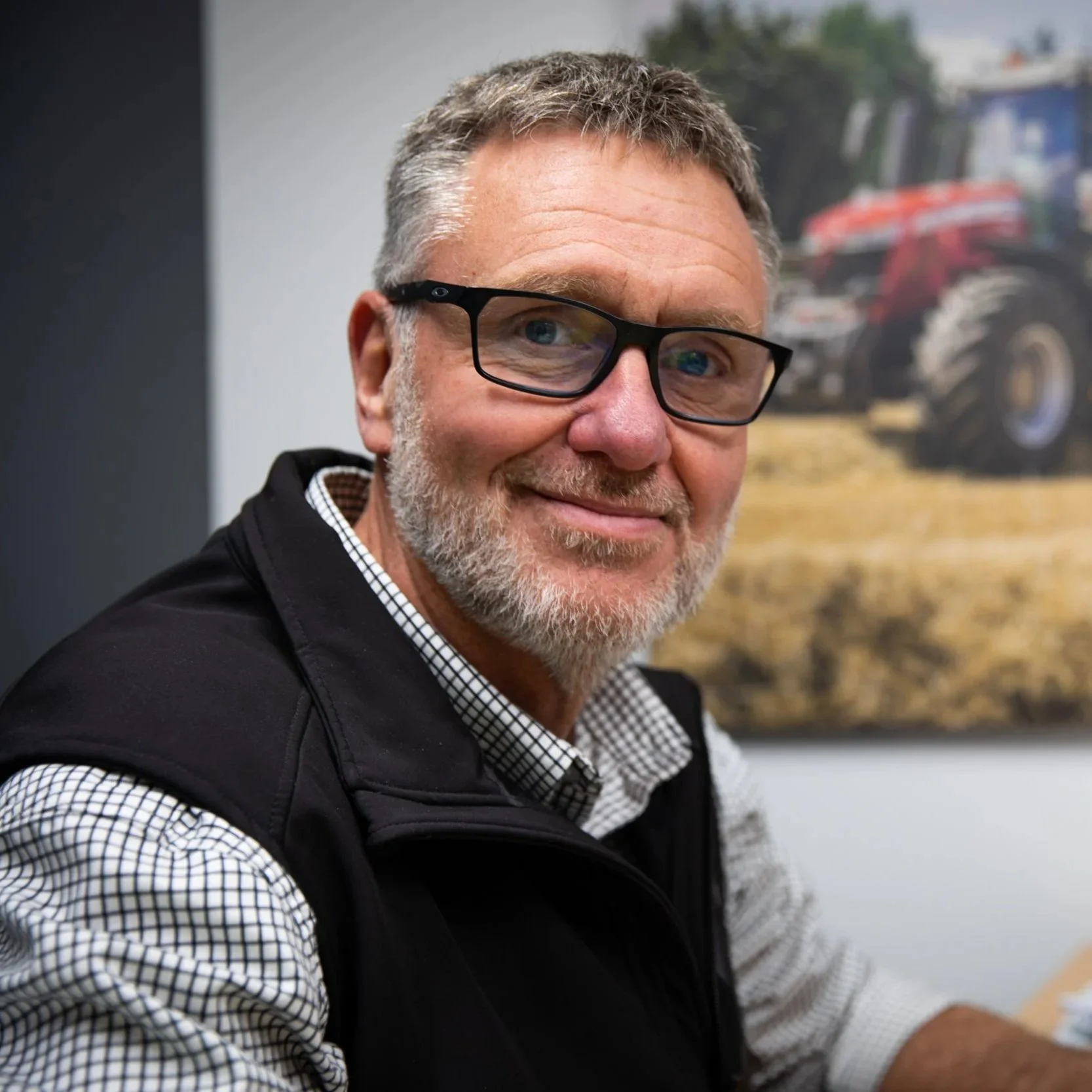 A man with gray hair and beard, wearing glasses, smiling, sitting indoors with a poster or picture of a tractor in the background.