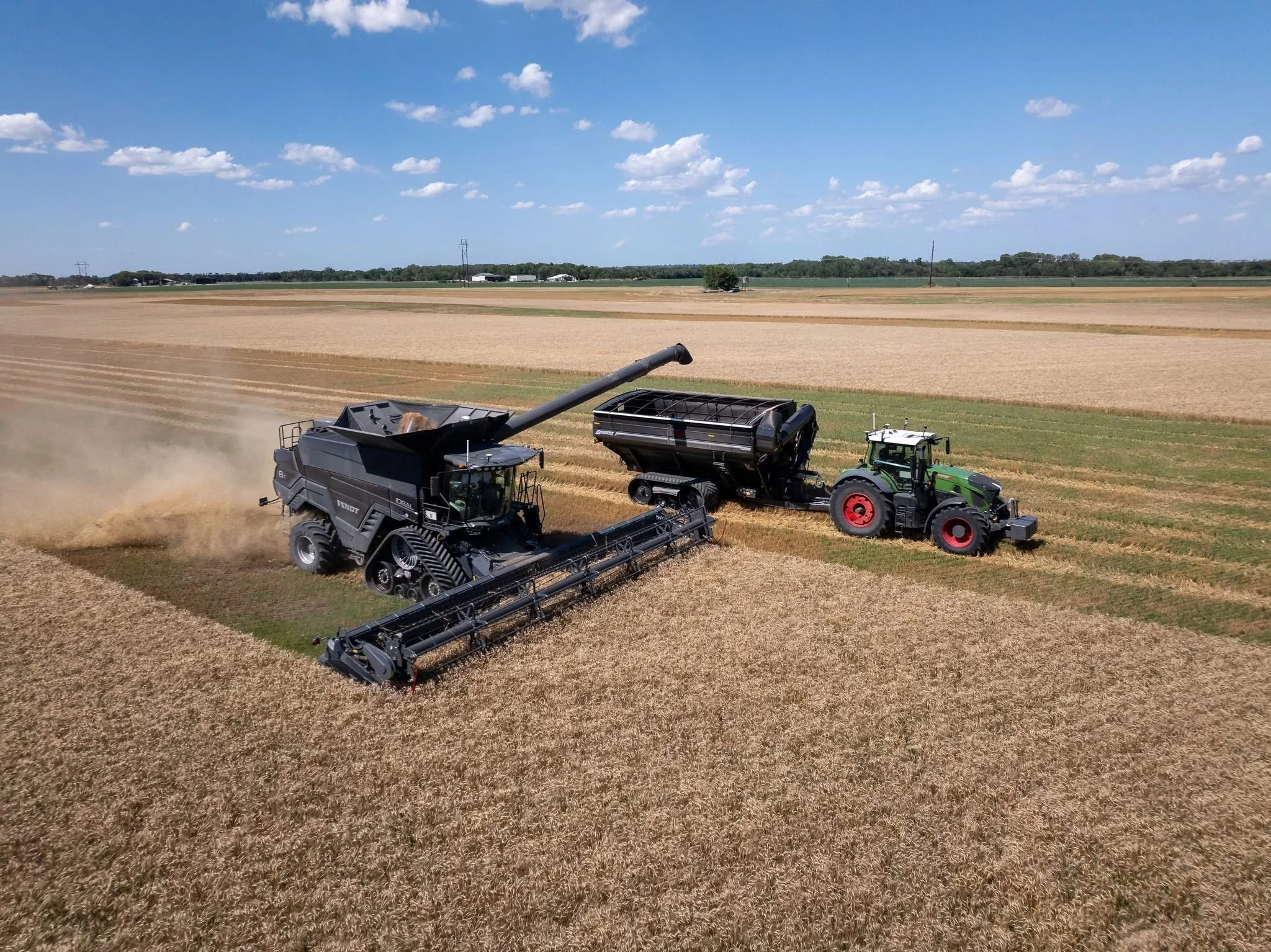 Two large farming machinery vehicles, one green and one black, working in a golden wheat field under a bright blue sky with scattered clouds.
