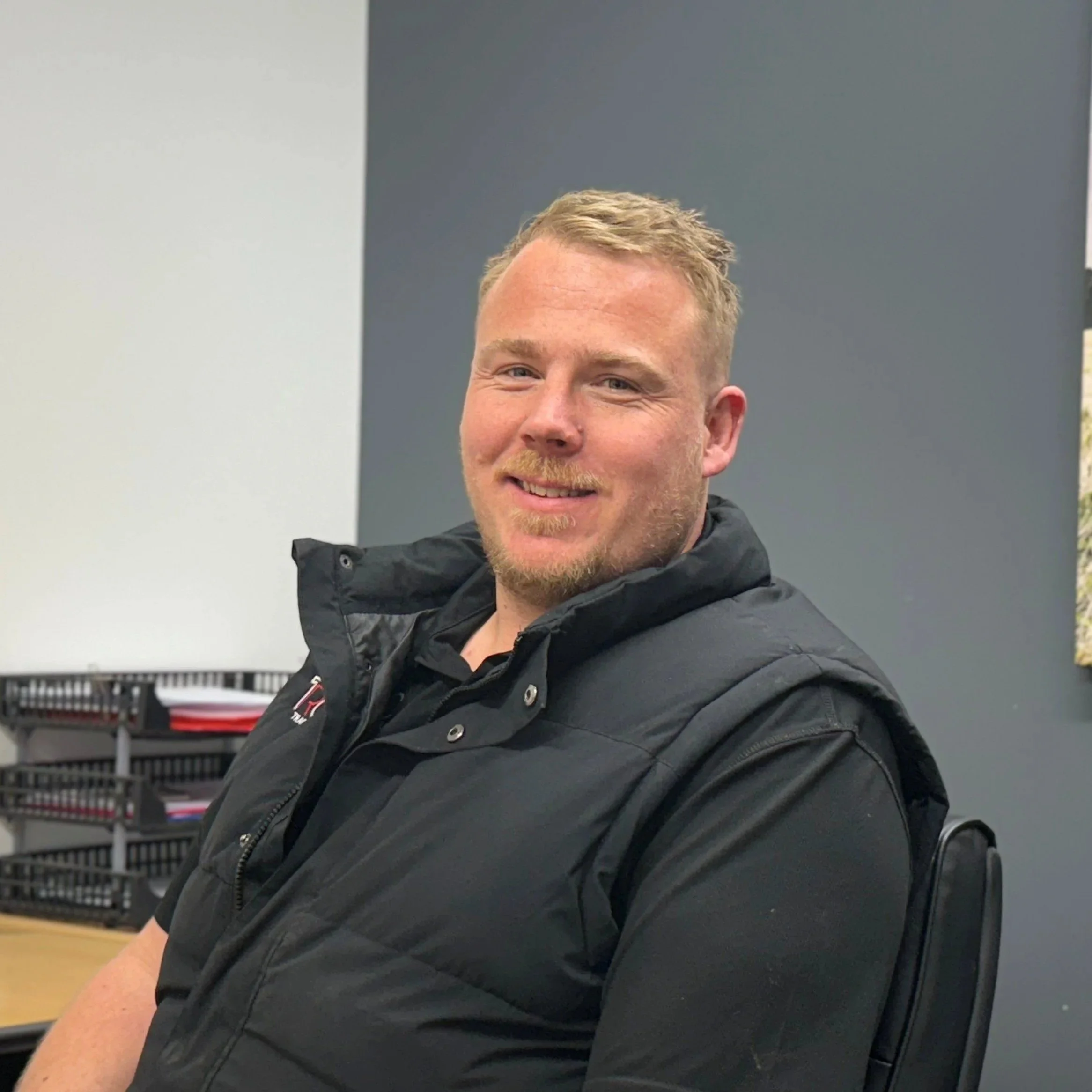 Close-up of a smiling man with blond hair and a beard, wearing a black jacket, sitting in an indoor setting.