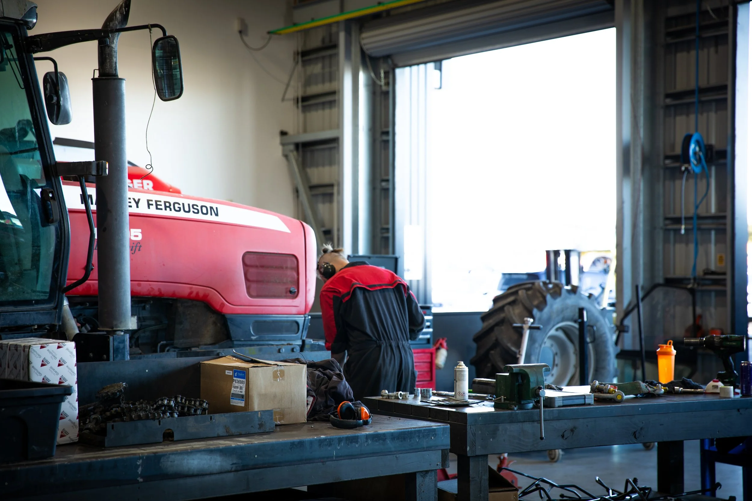Inside a workshop with a red tractor and various tools and parts on workbenches, a person wearing a red shirt and black jacket working at a desk, and an open garage door letting in daylight.