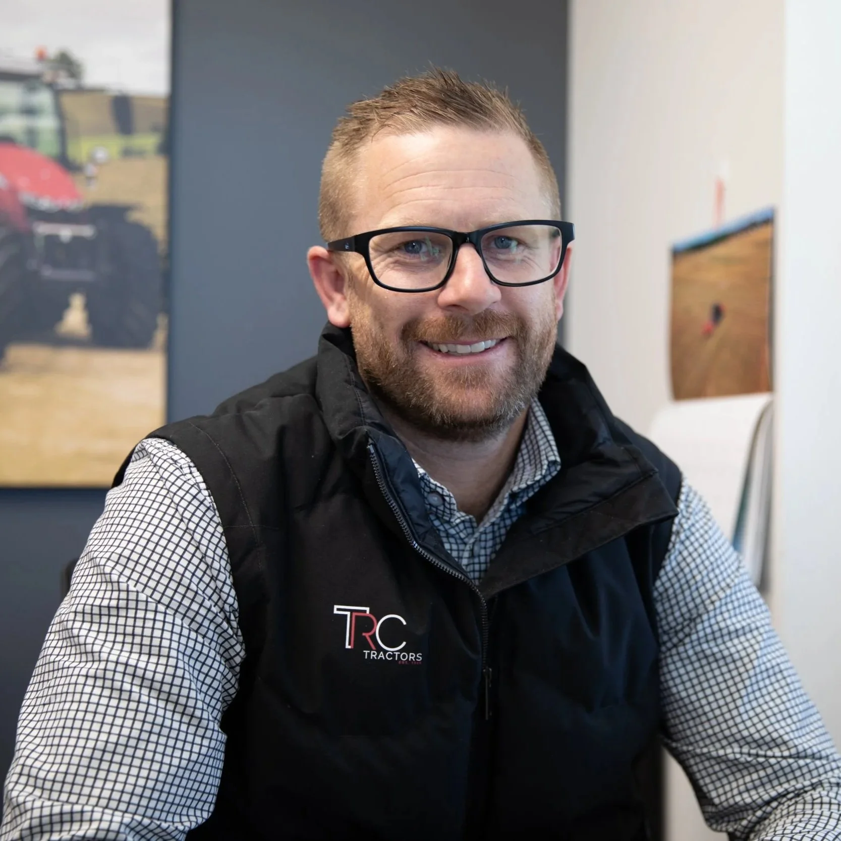 A smiling man with glasses and a beard, wearing a checkered shirt and a black vest with a logo that reads 'TRC Tractors,' sitting indoors with colorful posters or photographs in the background.