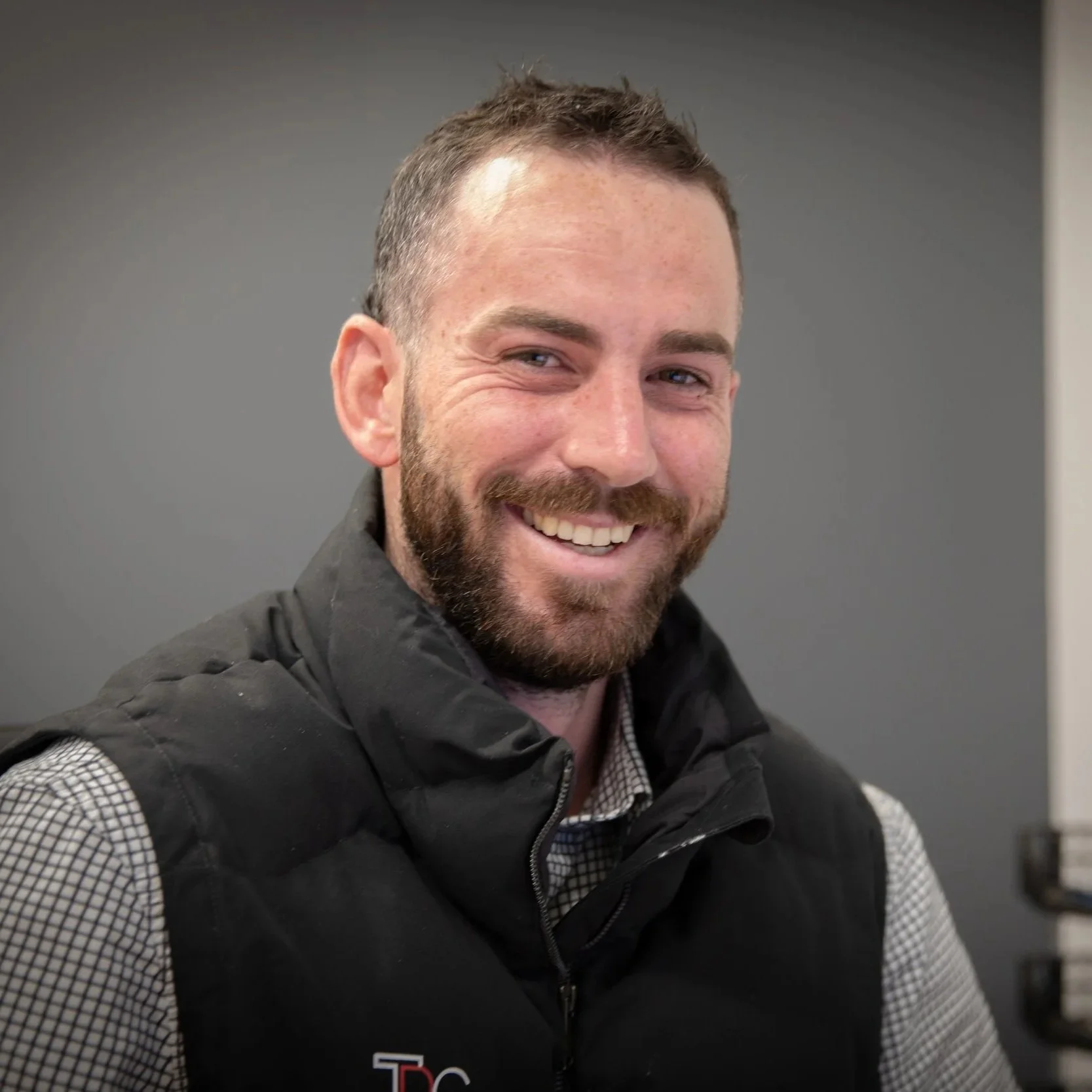 A smiling man with short hair and a beard, wearing a checkered shirt and black vest, sitting indoors against a gray wall.