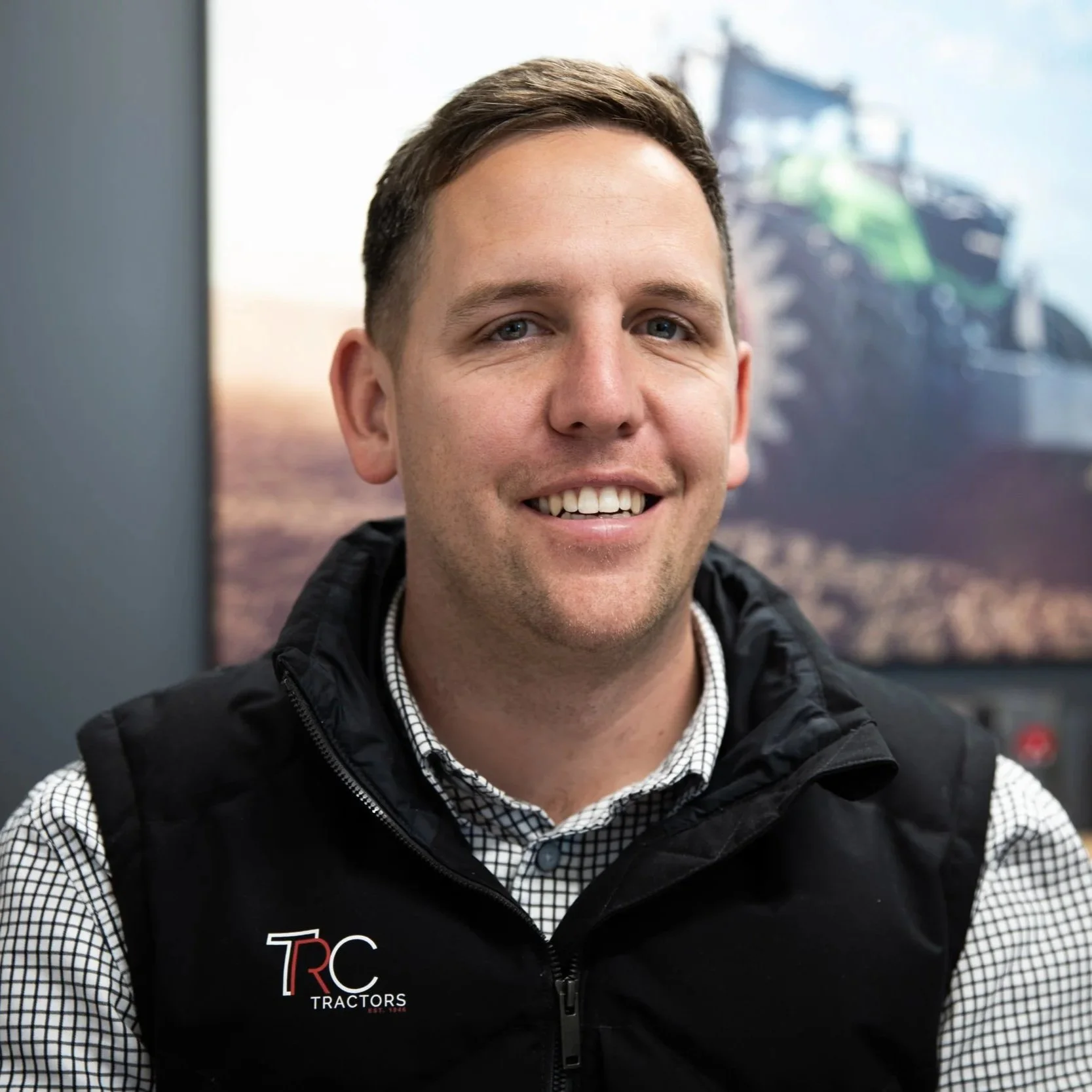 Close-up of a smiling man wearing a checkered shirt and a black vest with a logo, in front of a blurred outdoor background.