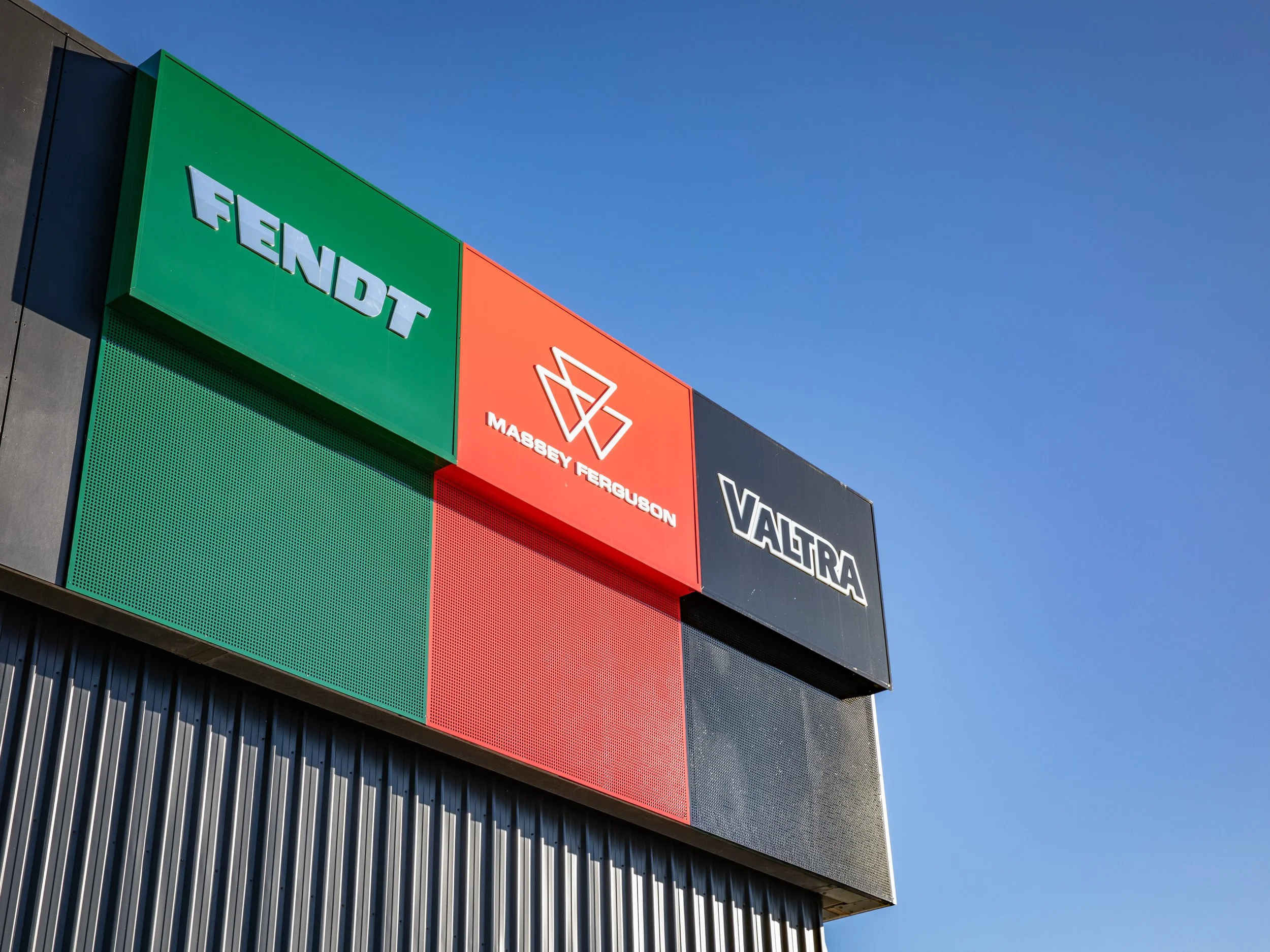 Close-up of building signage featuring logos of Fendt, Massey Ferguson, and Valtra against a clear blue sky.
