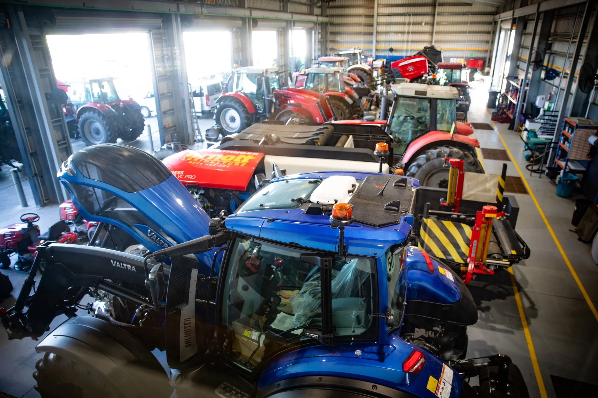 Inside a warehouse, multiple tractors and farm machinery are parked, including a blue tractor in the foreground, with tools and shelves on the walls.