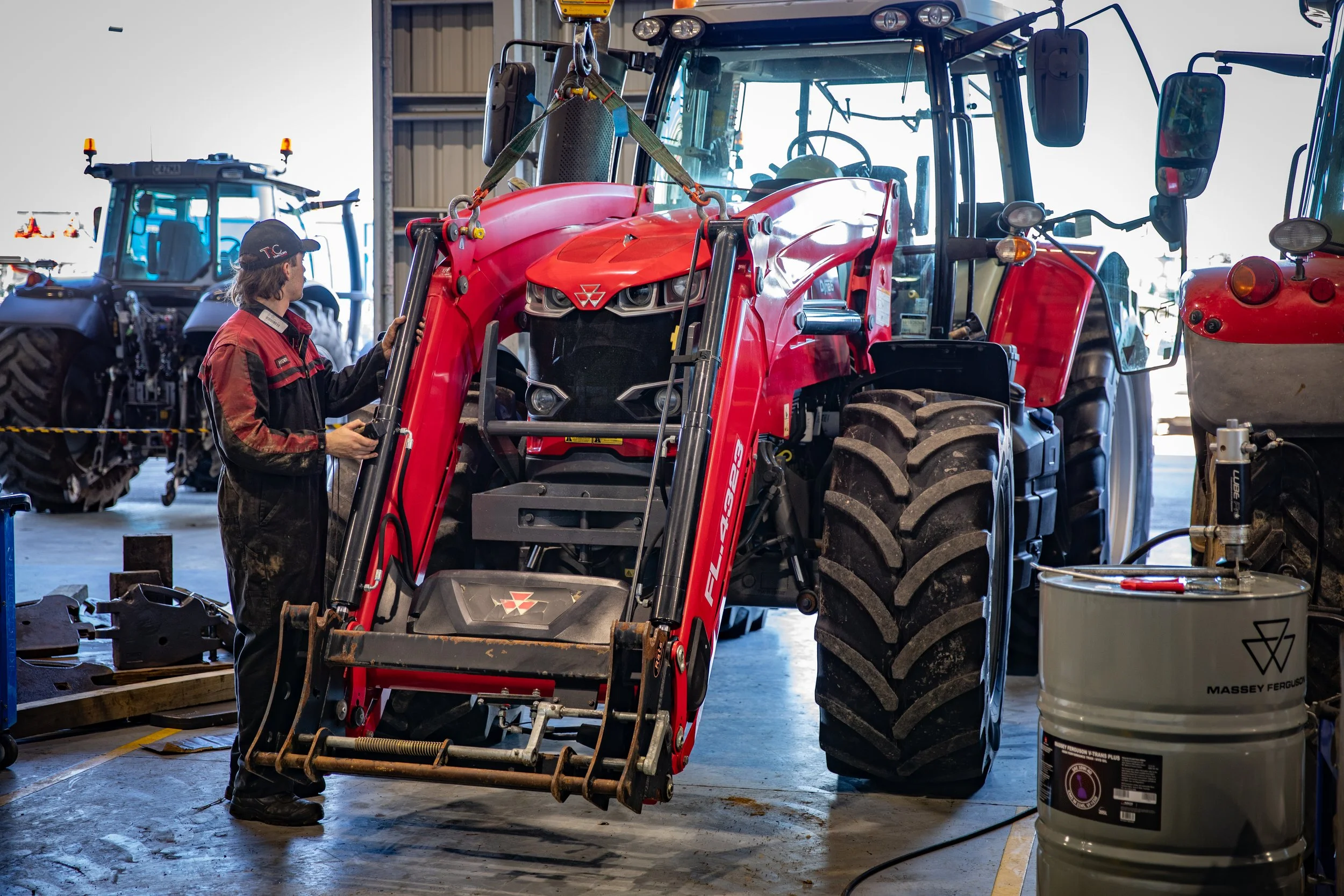 A person inspects a red tractor in a large indoor garage or workshop, surrounded by other tractors and equipment.