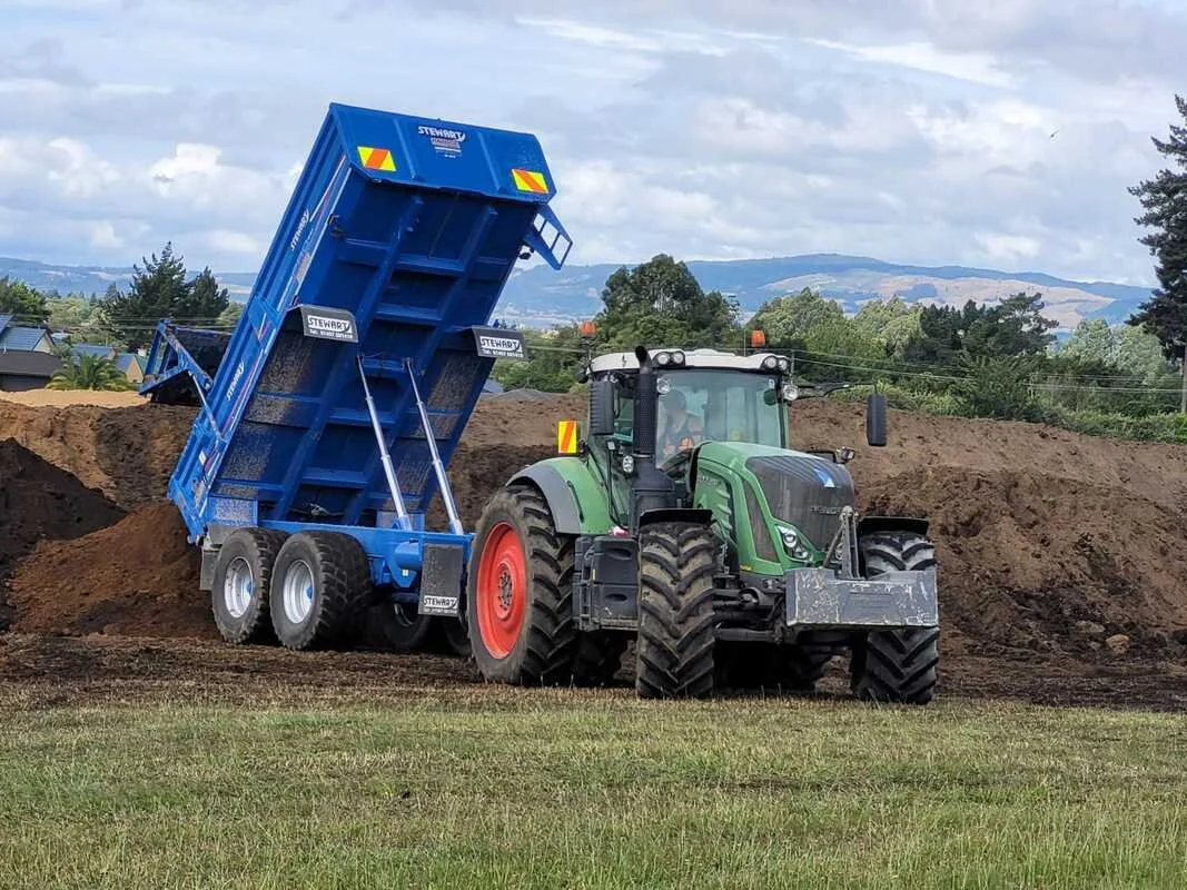 Green tractor with a grey front loader dumping dirt into a blue dump trailer at a construction site.