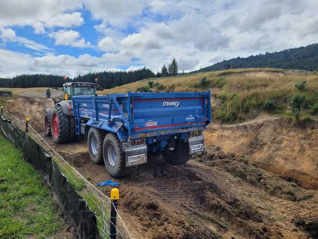 A blue tractor with a trailer attached is working on a farming site, moving dirt along a dirt path surrounded by green grass and a black fence with yellow posts, with rolling hills and a cloudy sky in the background.