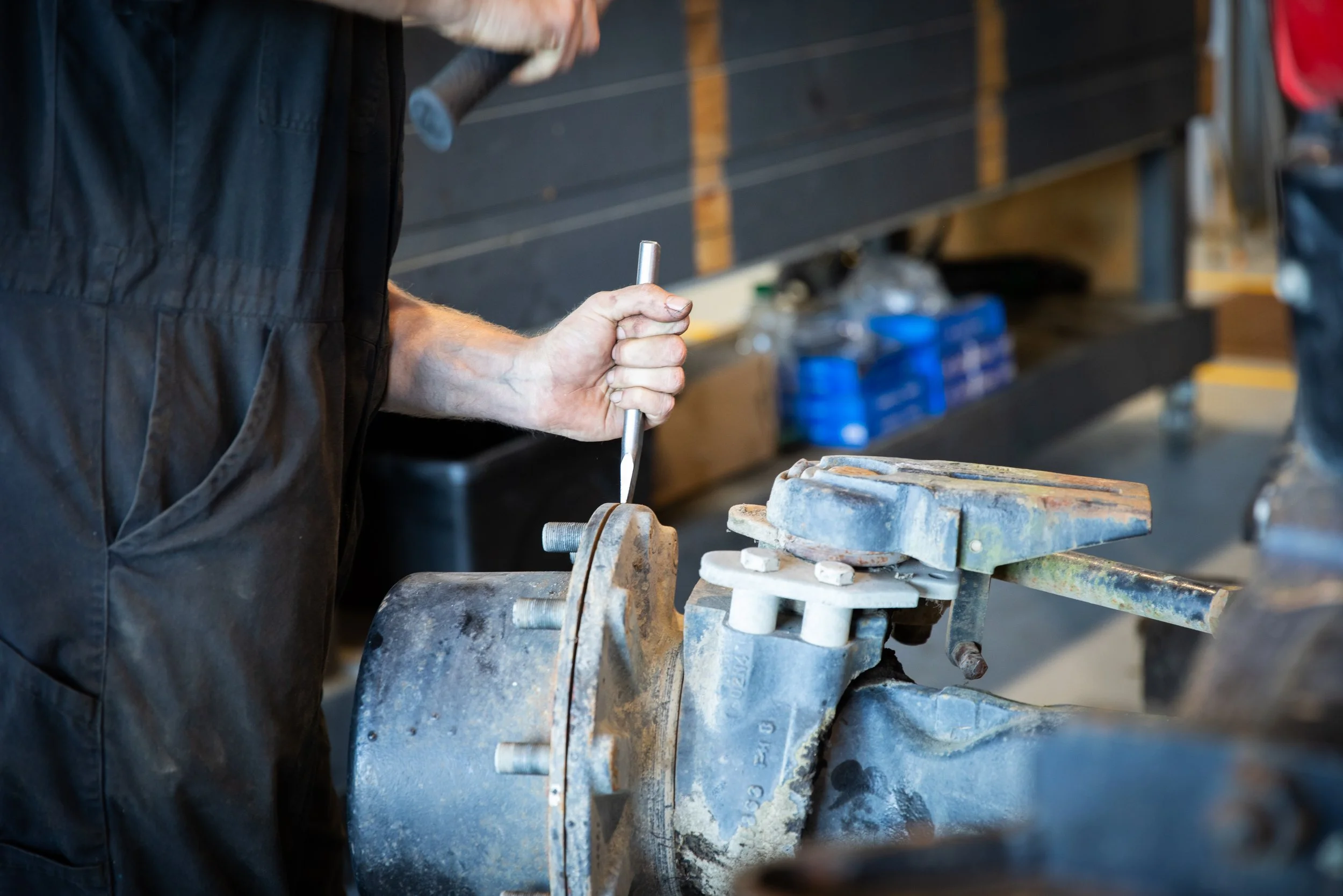 A person operating a metal lathe machine in a workshop, wearing a black work jacket and holding a tool while working on a piece of metal.