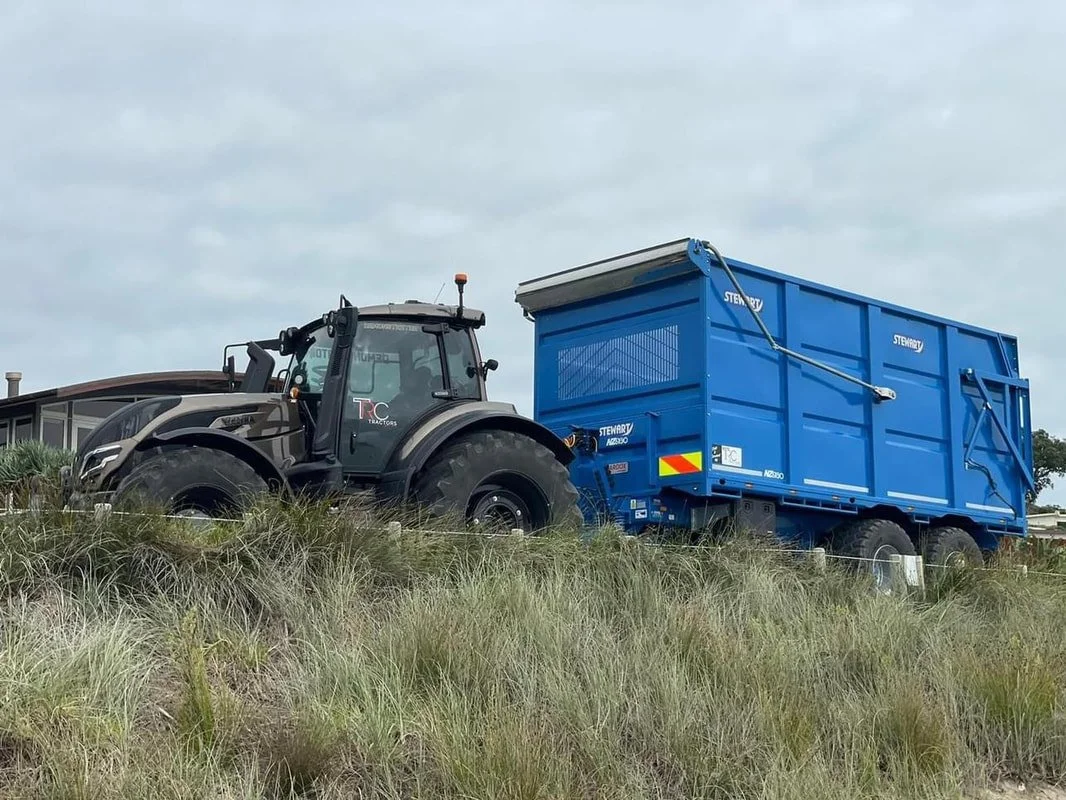A large black tractor with a blue container attached is parked on a grassy area under a cloudy sky.