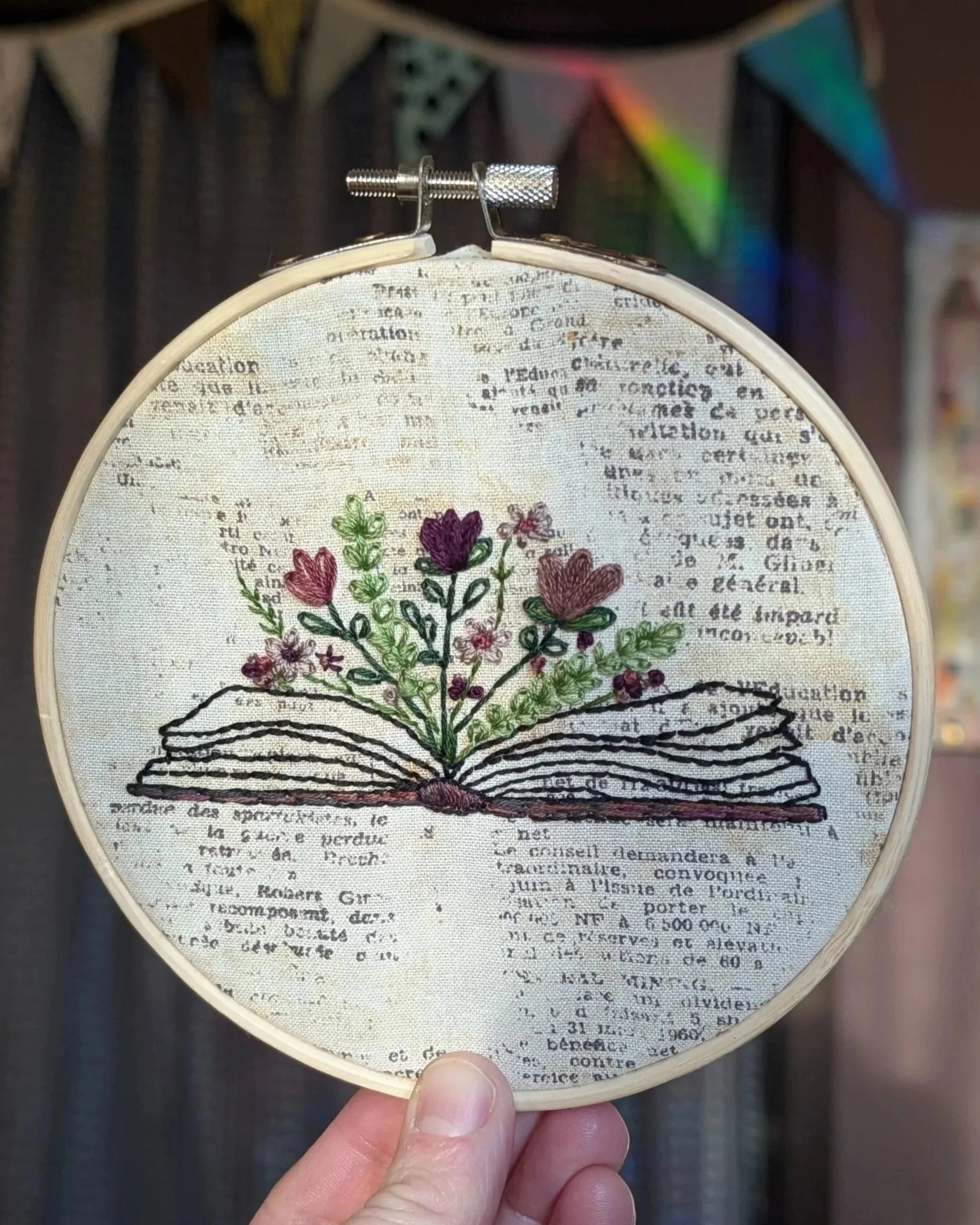 Close-up of an embroidery hoop with a detailed floral embroidery design on fabric, with the background showing colorful bunting flags.