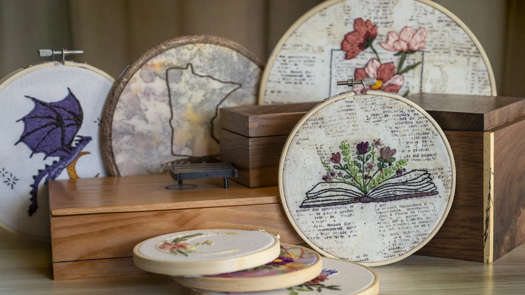 Hand embroidered hoops with floral and book-inspired designs displayed with handcrafted wooden boxes in a warm, minimal setting