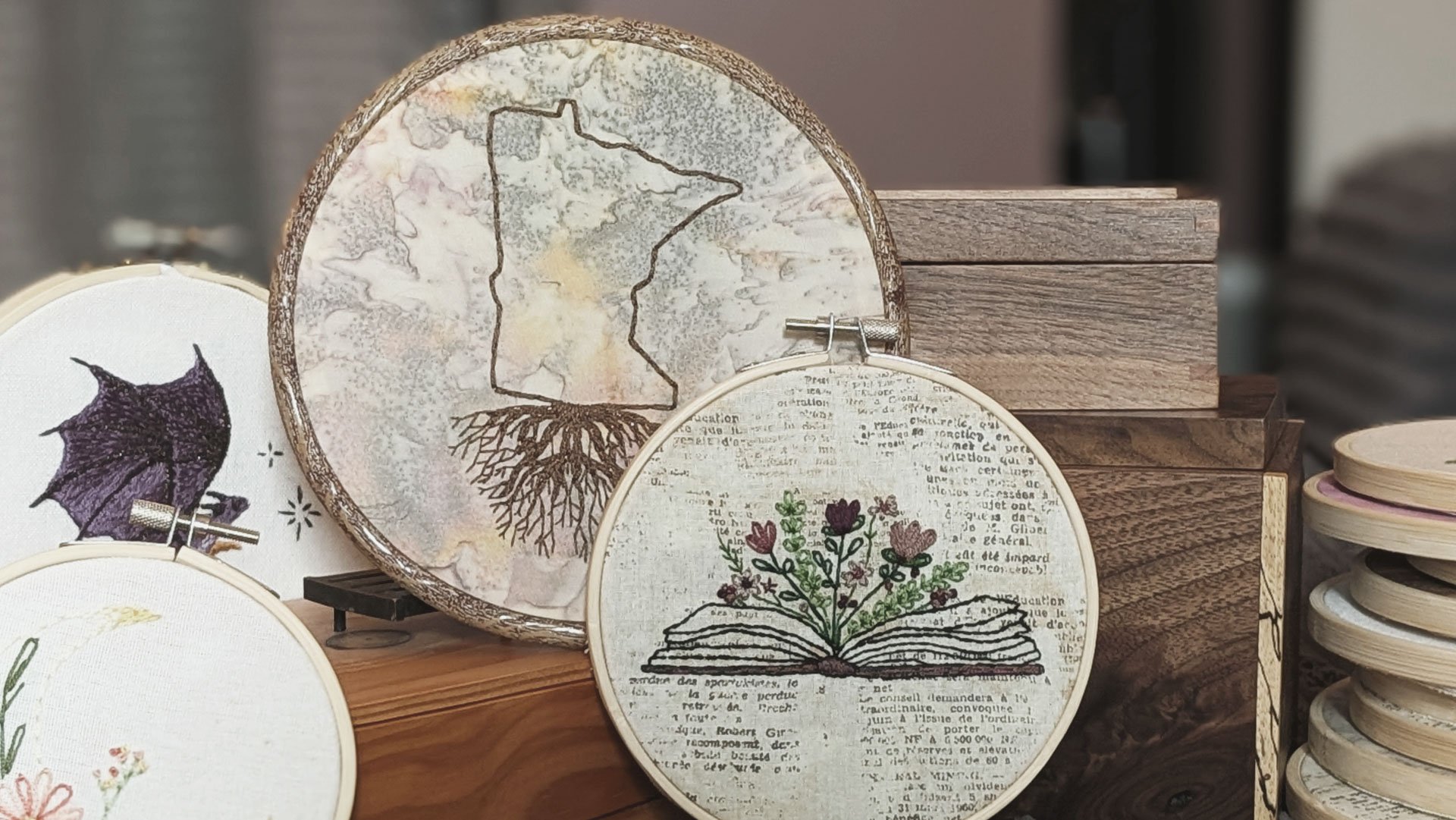 Various embroidered artwork displayed on a wooden surface, including a map of Minnesota with tree roots, a purple leaf, and a book with flowers growing out of it, among stacked wooden embroidery hoops.