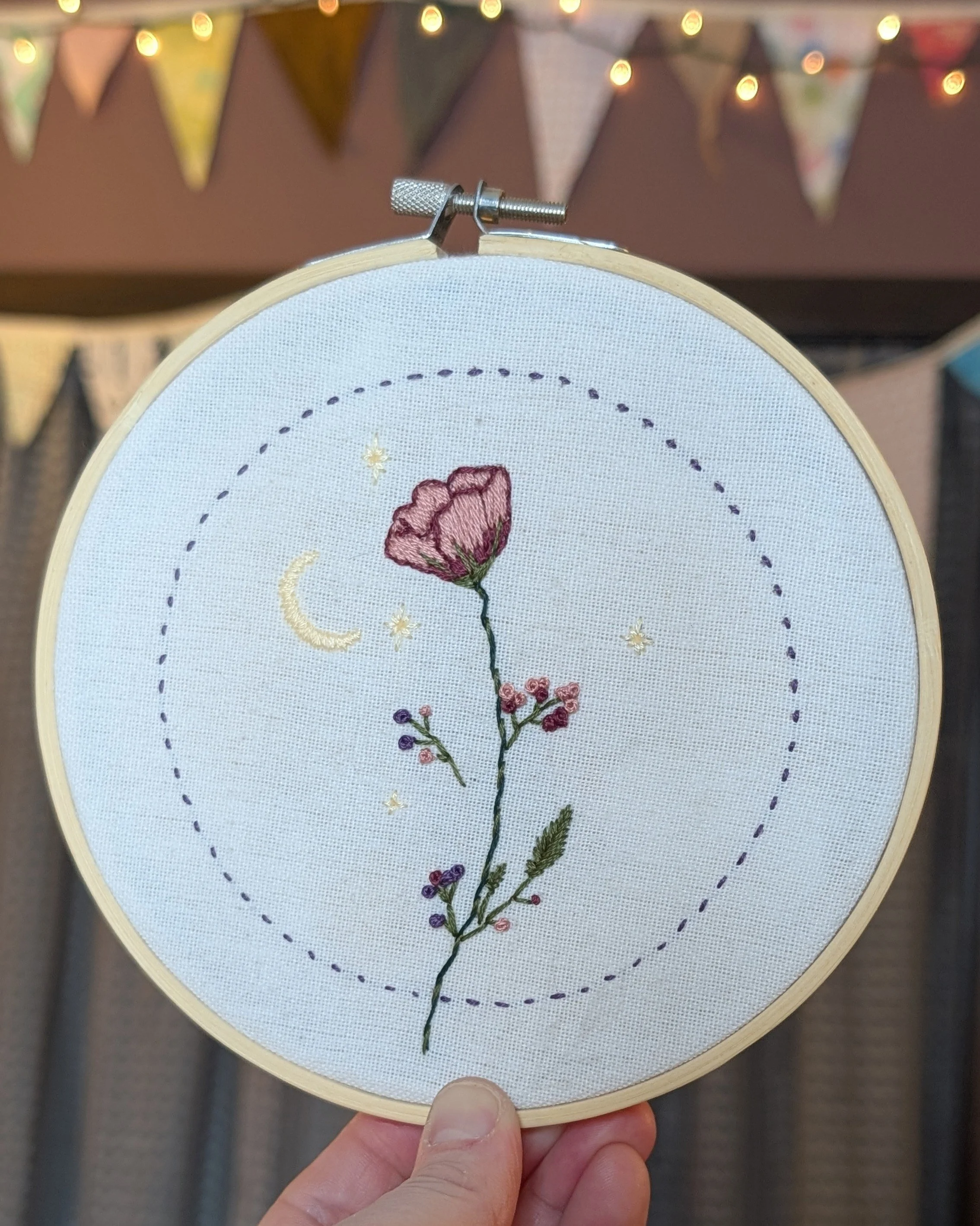 Close-up of a hand holding a small embroidery hoop with a floral embroidery design, featuring a pink flower and green stem, set against a decorated background with bunting and string lights.