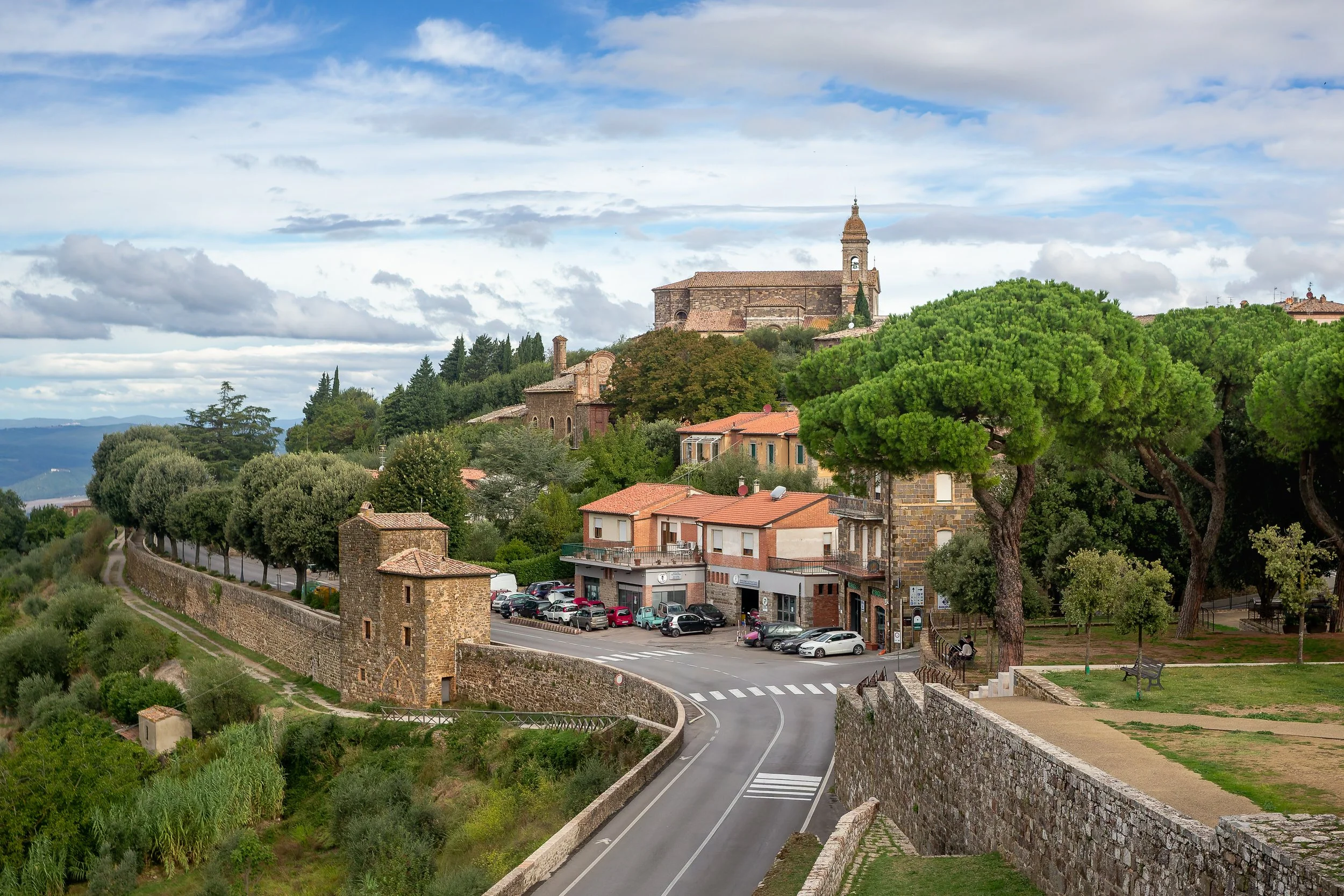 A scenic view of a small town with a historic stone church on a hilltop, surrounded by green trees, residential buildings, and a winding road.