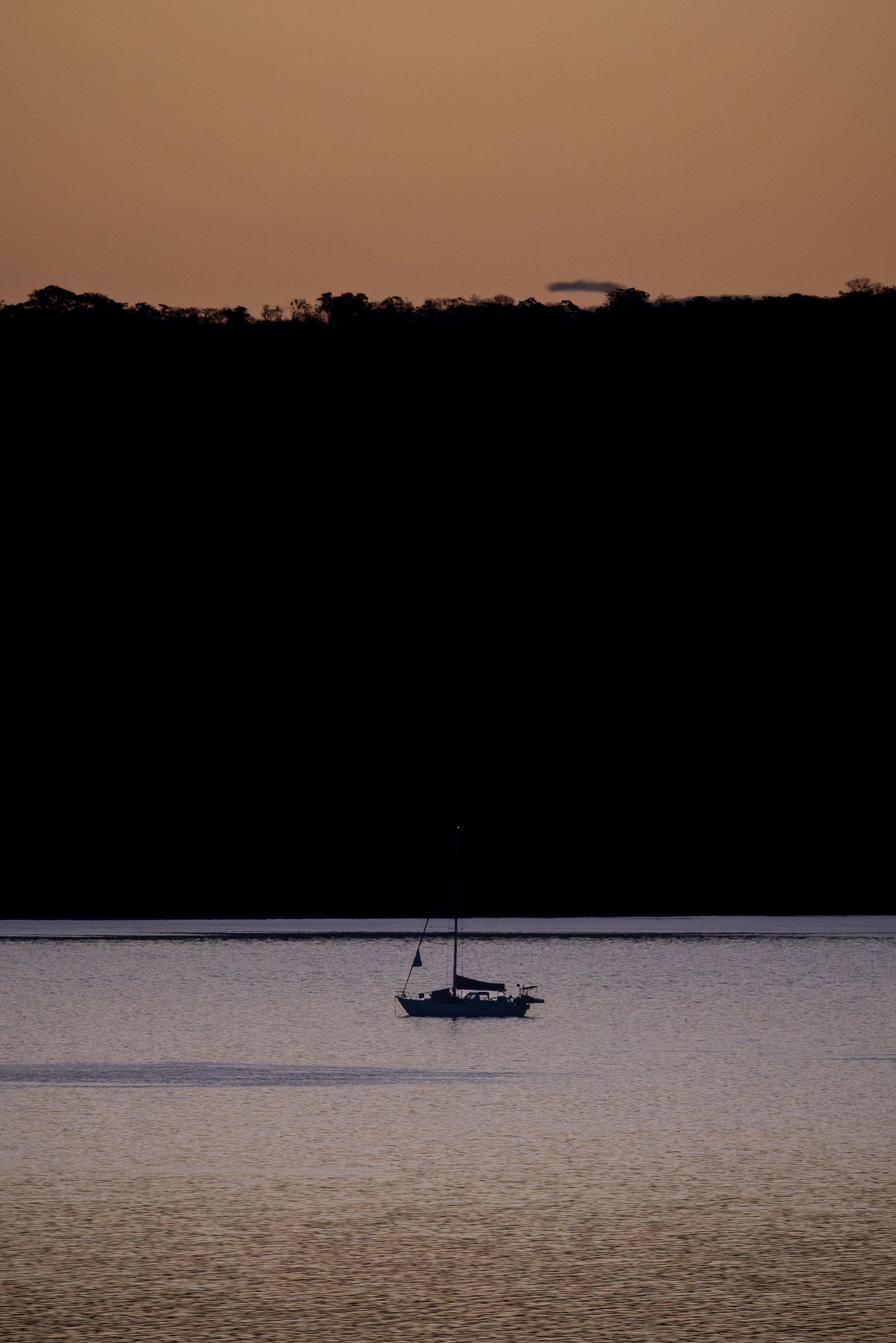 A sailboat on a calm body of water at sunset or dusk with a silhouetted tree line in the background.