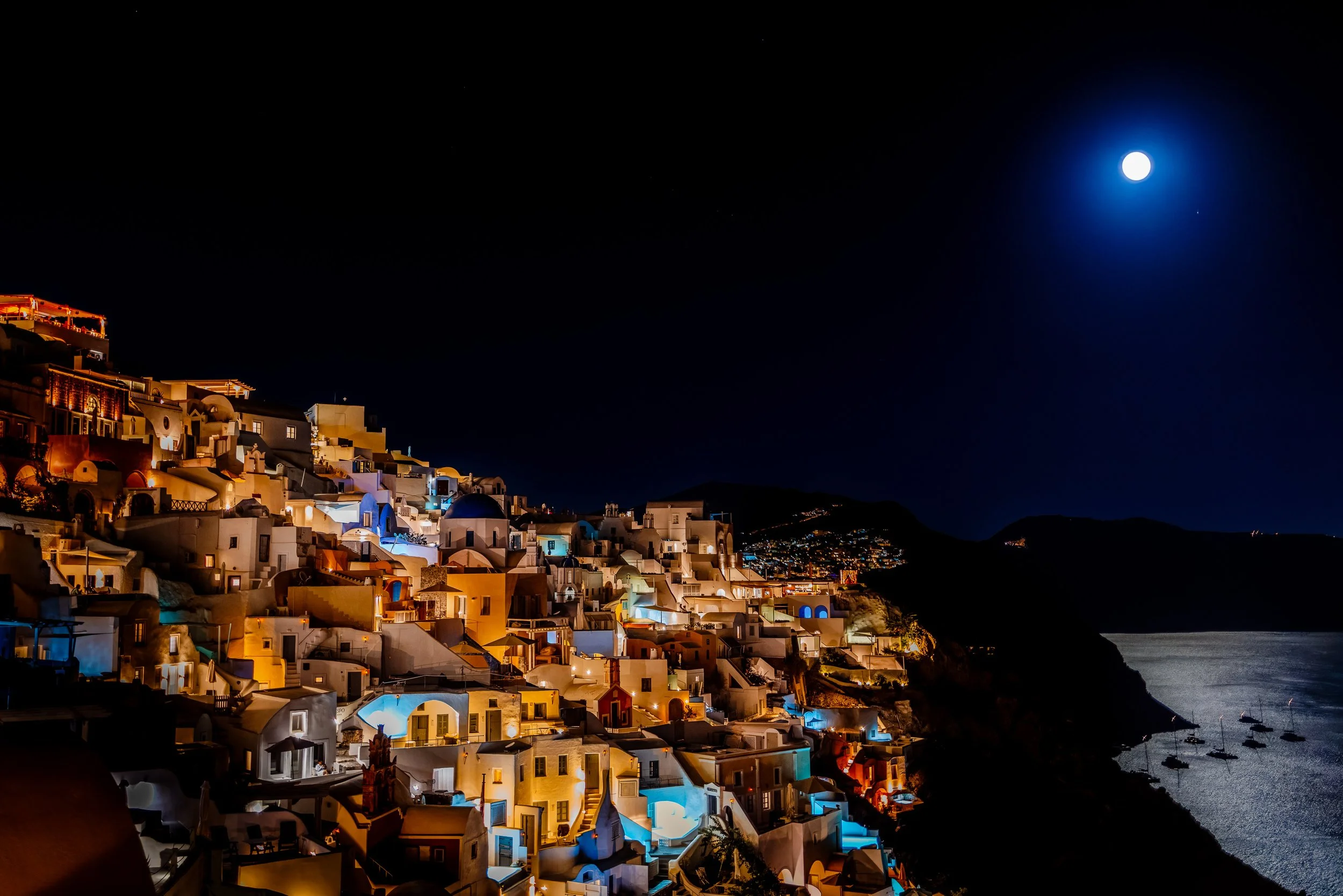 Moonlit night view of the illuminated cliffside village of Oia overlooking the Aegean Sea on Santorini island, Greece.